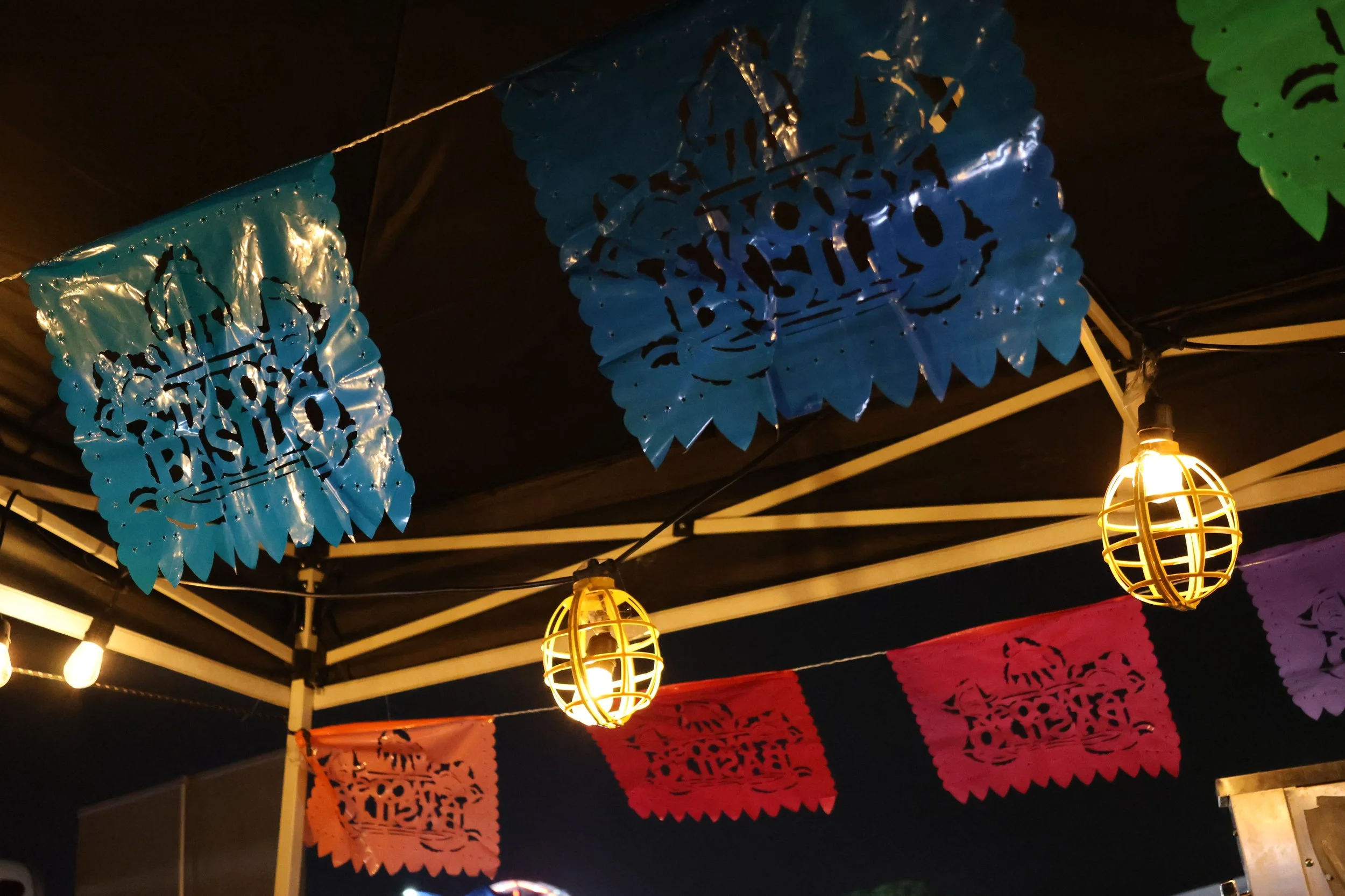 Decorative papel picado banners in blue, pink, and orange hanging from a dark ceiling, illuminated by hanging lantern lights, likely for a celebration or festival.