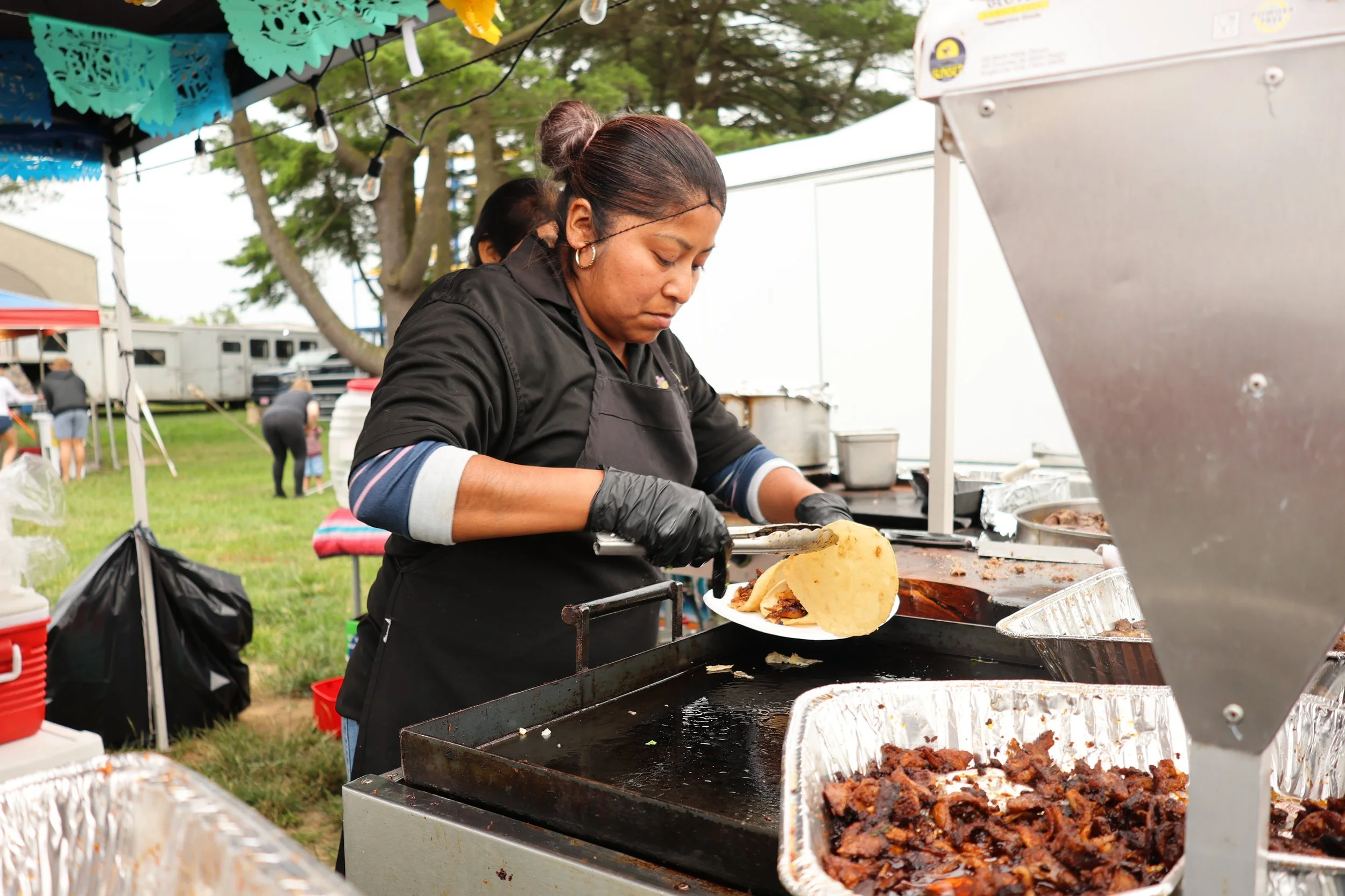 A woman preparing Mexican food at an outdoor food stall, serving a taco filled with meat and toppings.