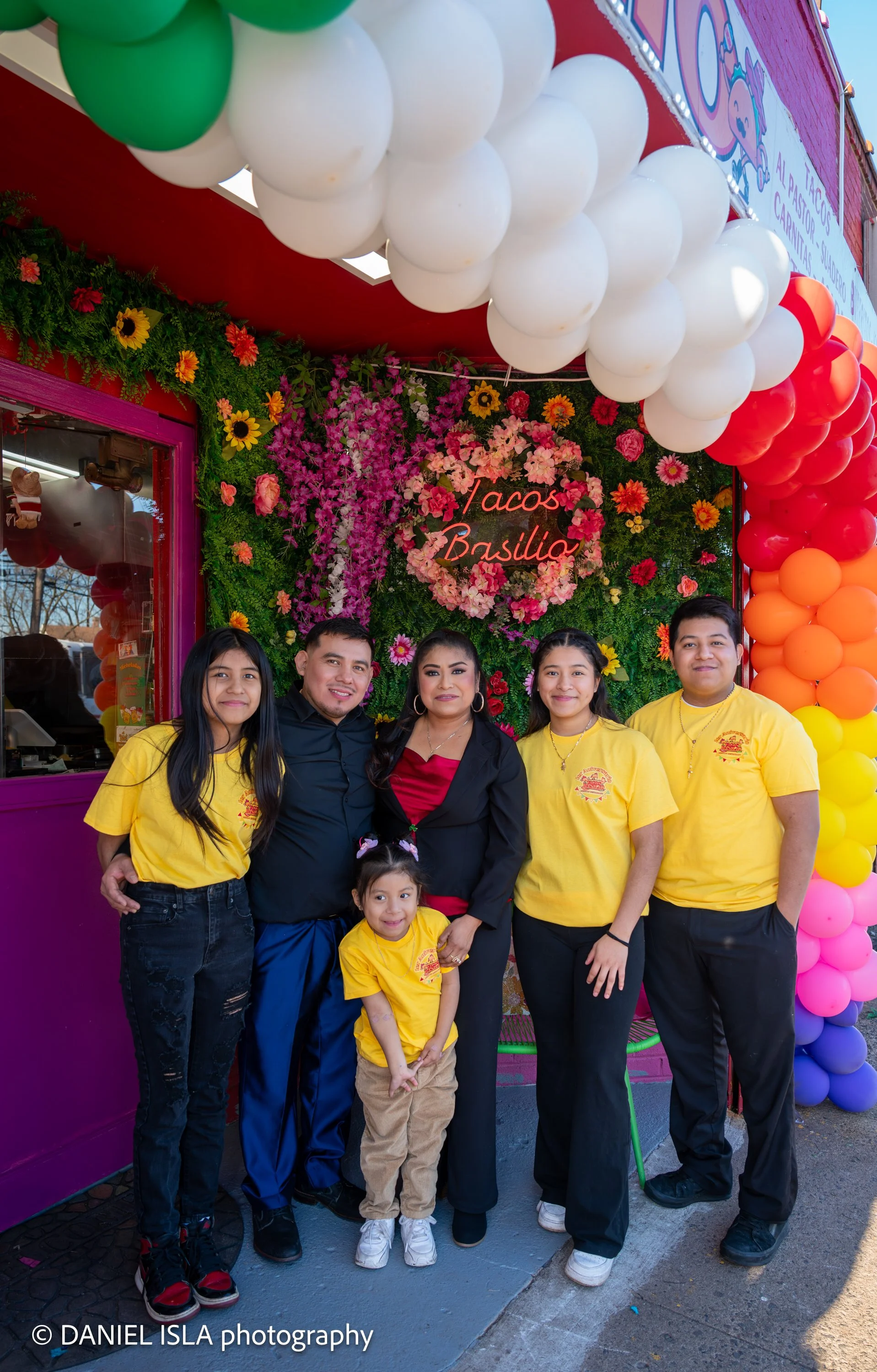A group of five people, four women and one man, standing in front of a decorated wall with flowers and a sign that reads 'Tacos Basilio'. They are smiling and wearing casual clothing, with some women wearing yellow shirts and a young girl in front we