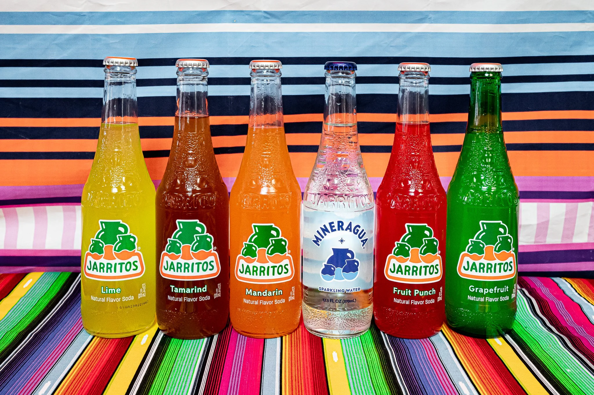 Six glass soda bottles with metal caps on a colorful striped tablecloth. The bottles are of different flavors: lime, tamarind, mandarin, sparkling water, fruit punch, and grapefruit. The bottles are placed in front of a blue, black, and orange stripe