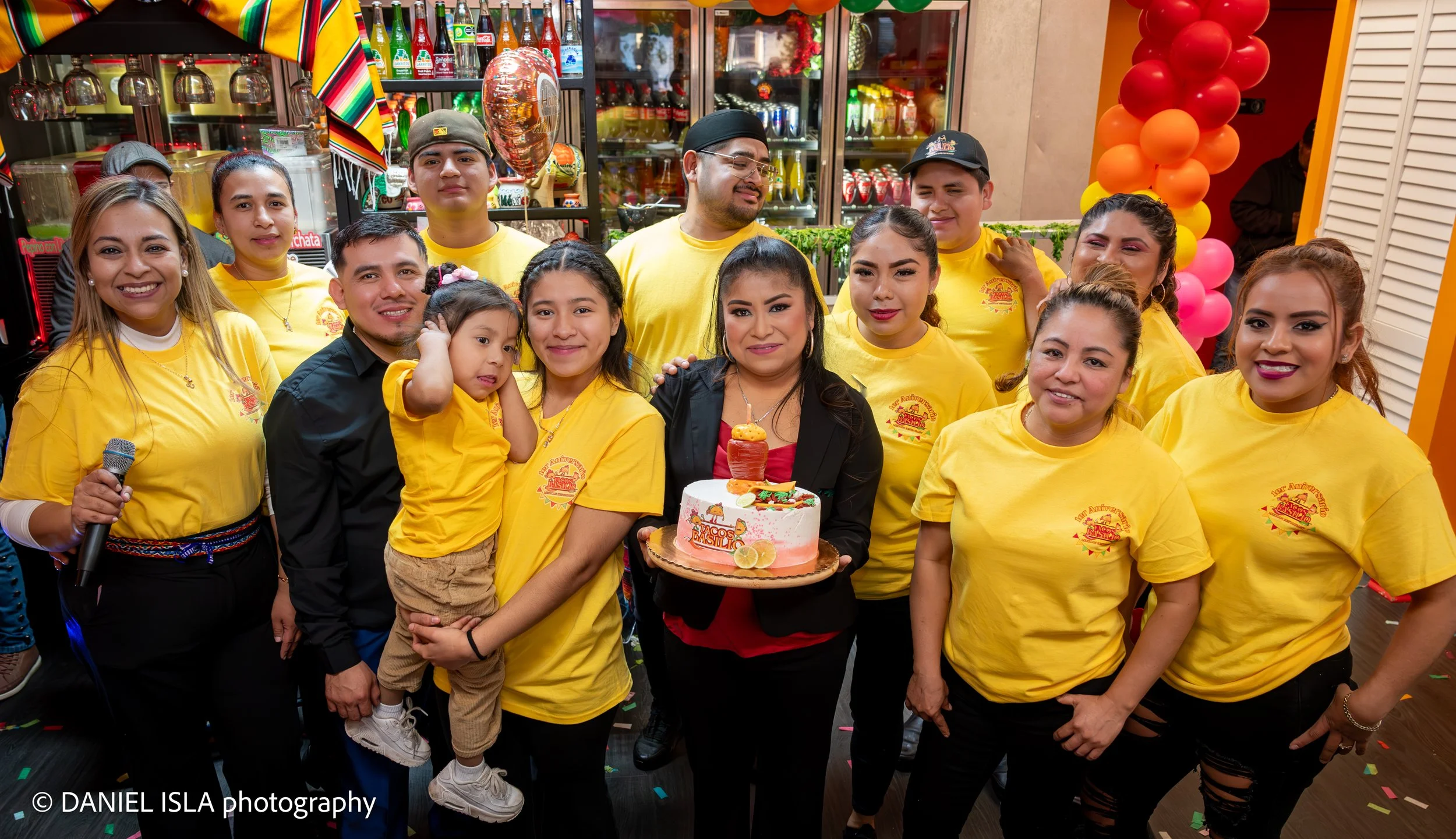 A group of people celebrating a birthday party, with many smiling and posing for the photo. Some are wearing yellow shirts with a logo, and a woman in the middle holds a cake with decorative toppings. The background features a decorated space with ba