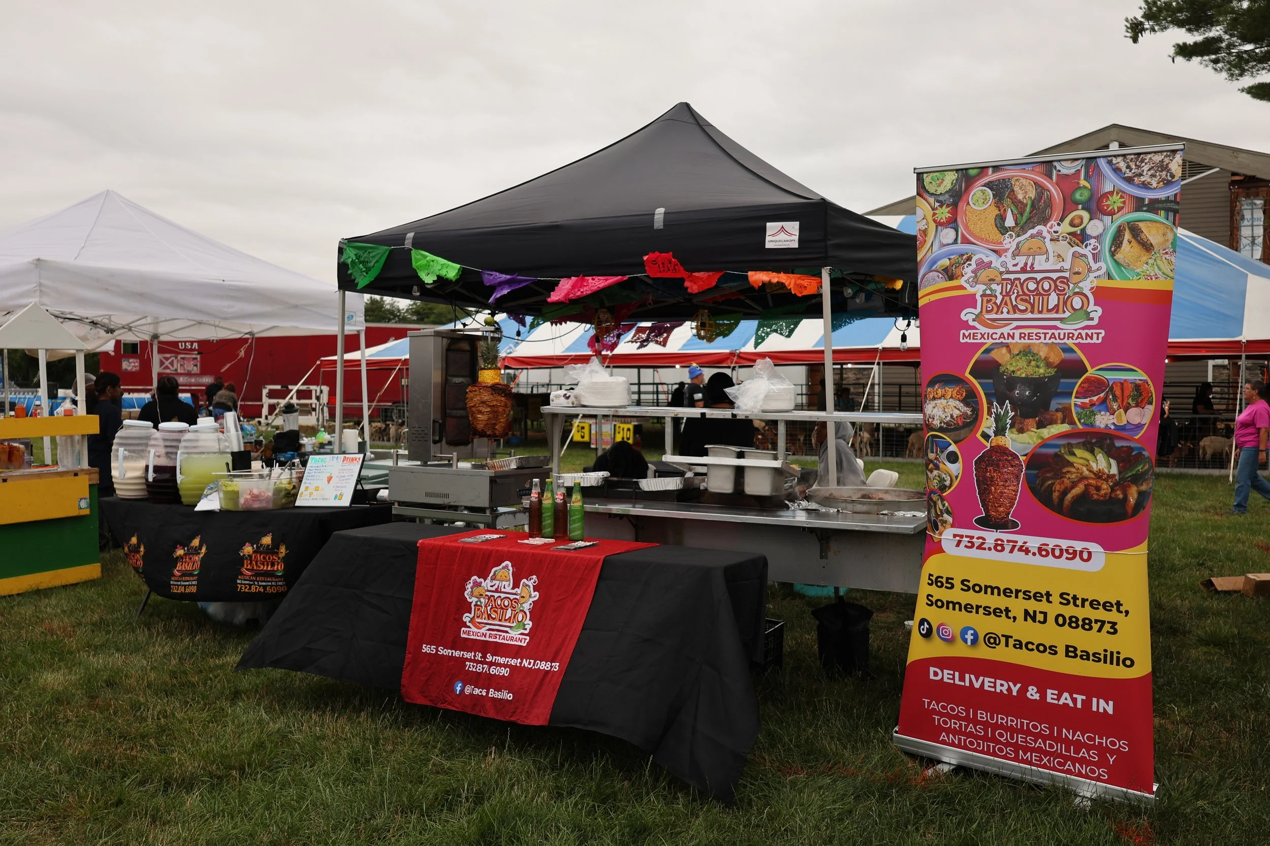 Outdoor food stand at a festival, featuring a black canopy with colorful papel picado banners, a promotional banner for Tacos Basilio, and various food preparation and serving items on tables, with people in the background.