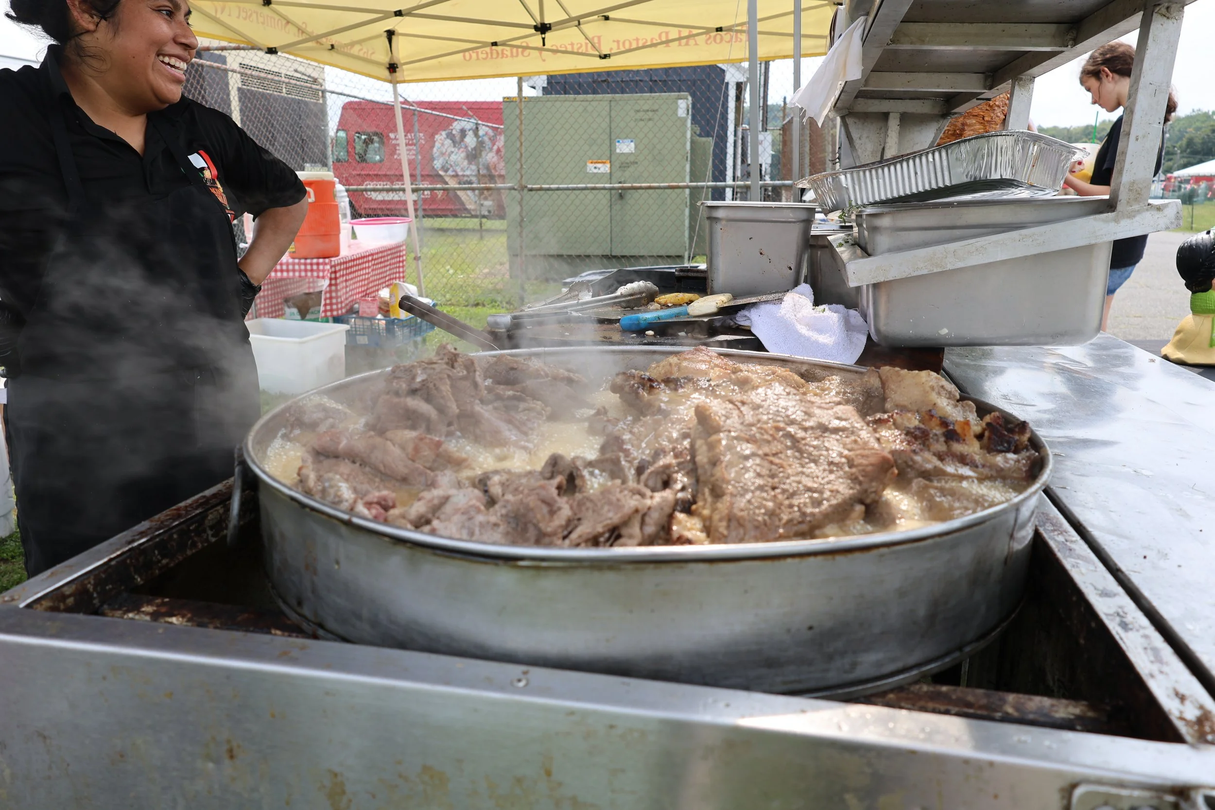 A woman cooking meat on a large outdoor grill at a food stand with a yellow tent overhead, people in the background, and a chain-link fence surrounding the area.