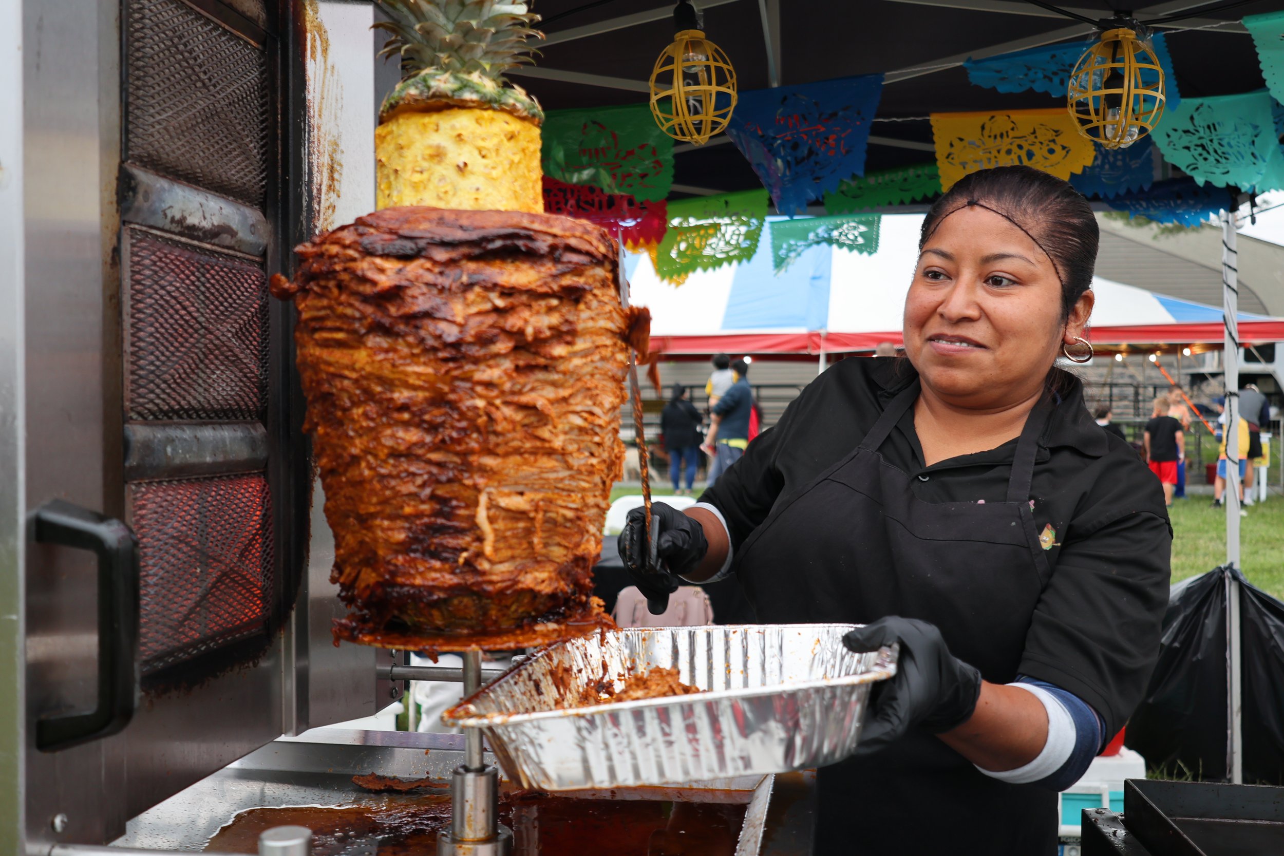 A woman preparing a traditional Mexican dish called birria, with a vertical spit of roasted meat, at an outdoor fair or festival. The woman wears black gloves and is serving the meat into a tray, with colorful decorations and other people visible in 