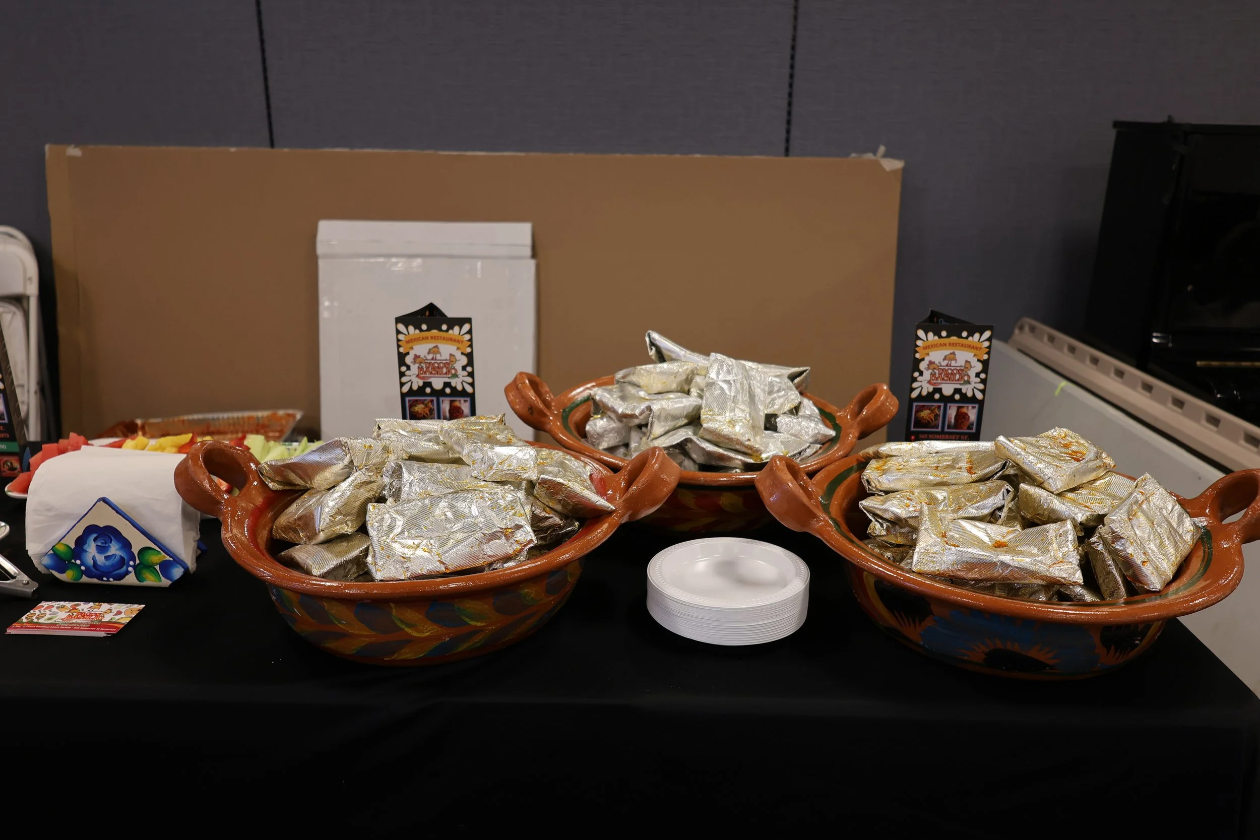 Two large traditional Mexican pottery bowls filled with foil-wrapped items, likely tamales, on a black table. There are also small paper plates, a small box of tissues, a stack of napkins, and promotional cards for a Mexican restaurant on the table. 