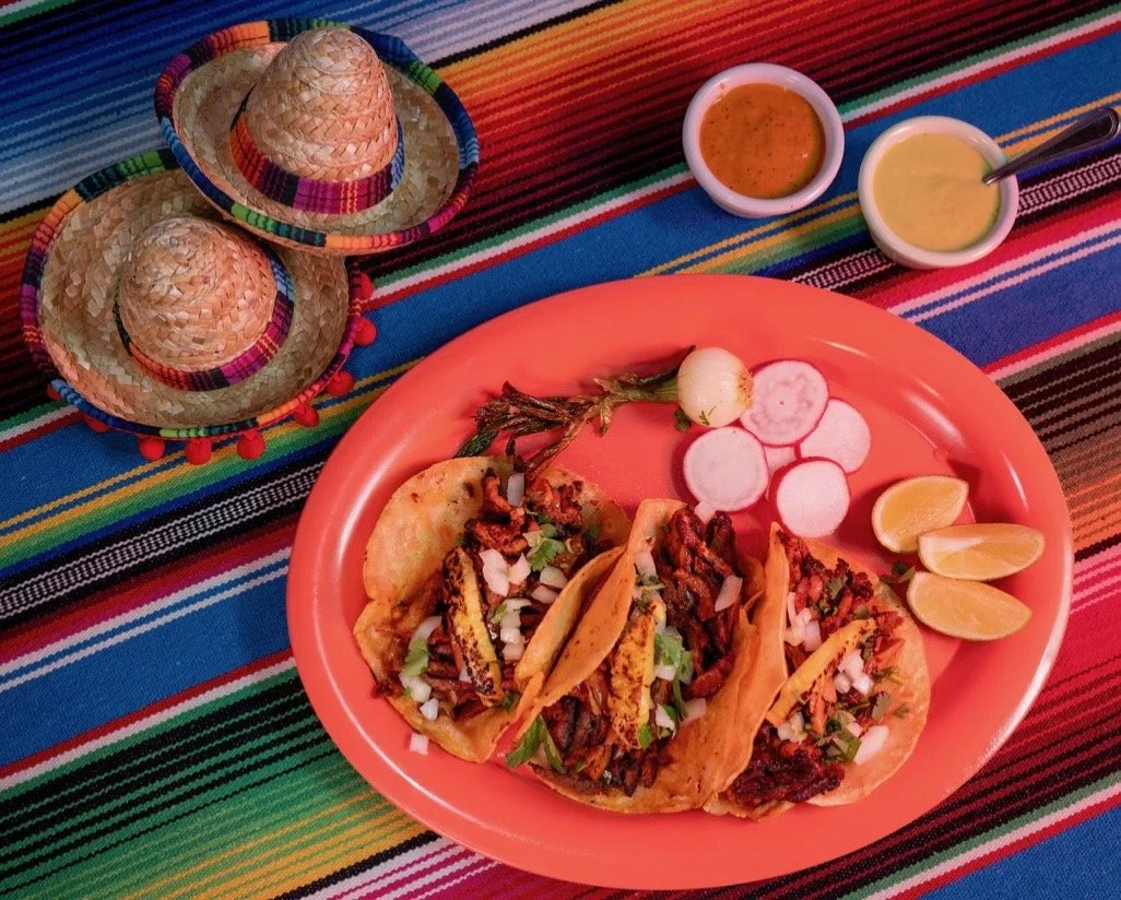 Three tacos with grilled meat, onions, and herbs on an orange plate, accompanied by radish slices, lime wedges, and a small onion, with salsa and sauces on the side, all on a colorful striped tablecloth with two small sombreros.