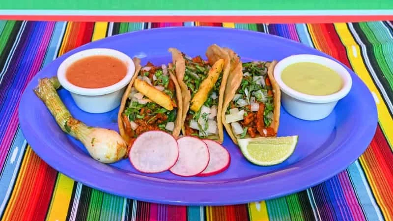 Three tacos filled with chopped meat and vegetables on a blue plate, with sliced radish, lime wedge, and sauces on the side, on a colorful striped tablecloth.