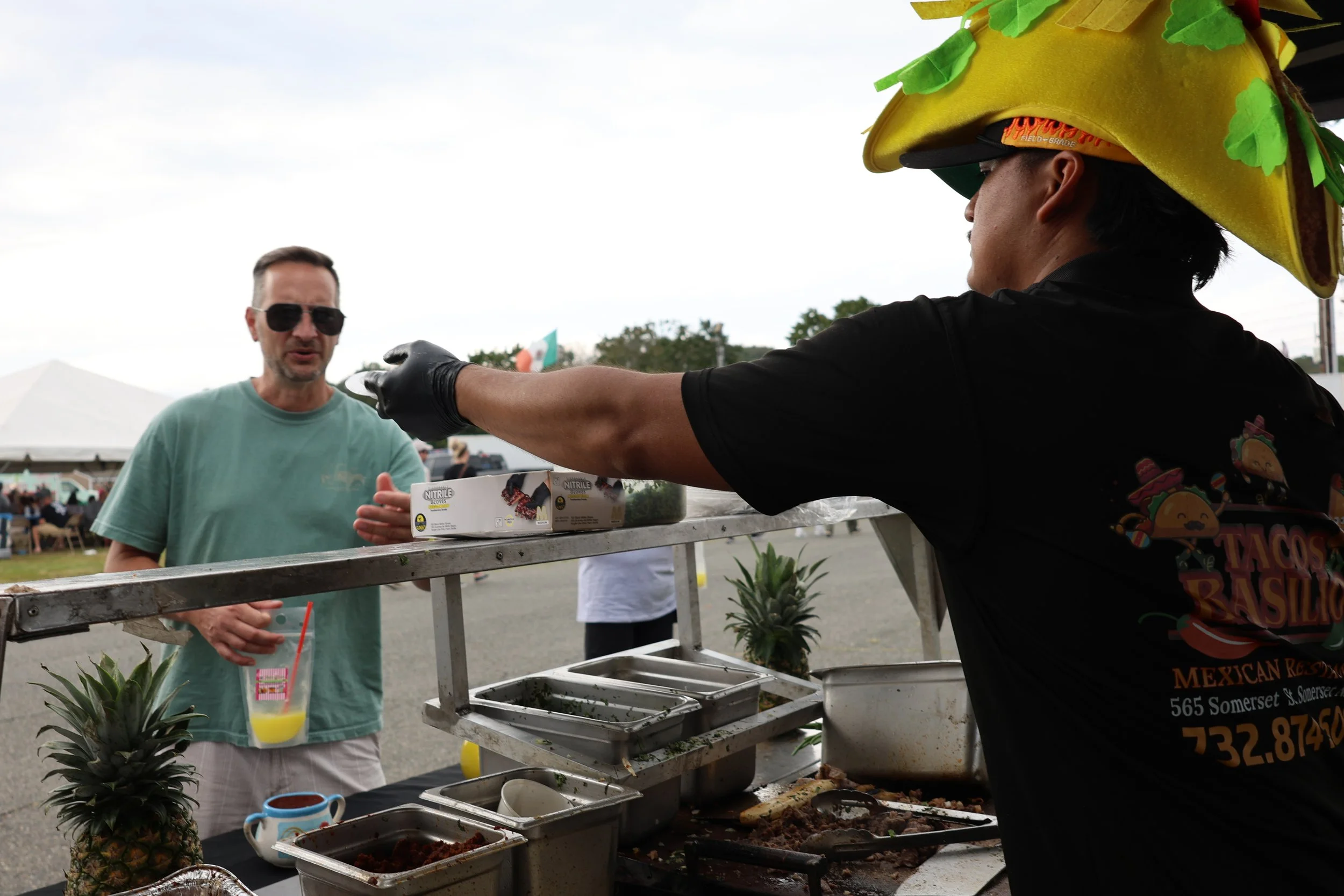 A man in a yellow taco-shaped hat at a food stand, serving food to a customer. The customer is wearing sunglasses and a teal shirt, holding a drink with a straw. Pineapples are on the table, along with various food containers.