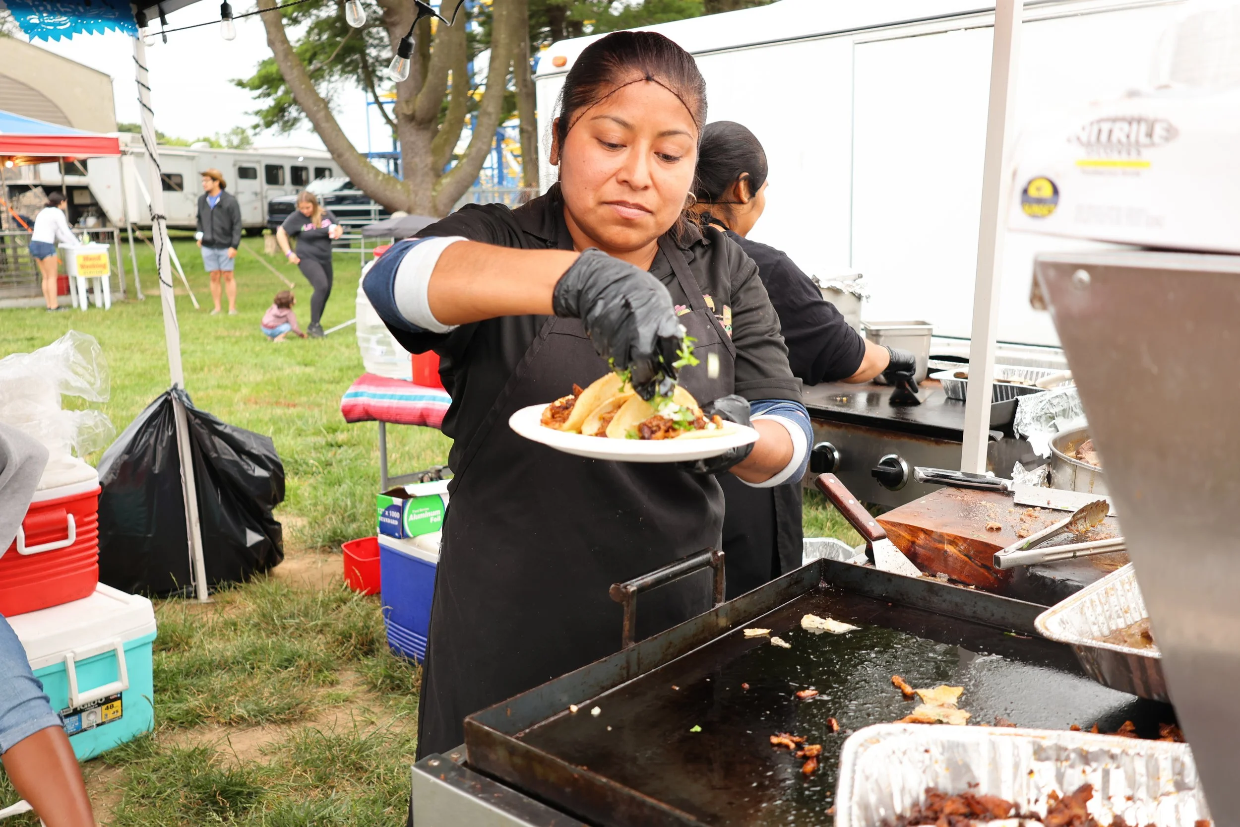 Woman serving tacos at an outdoor event with people in the background, trees, and tents.