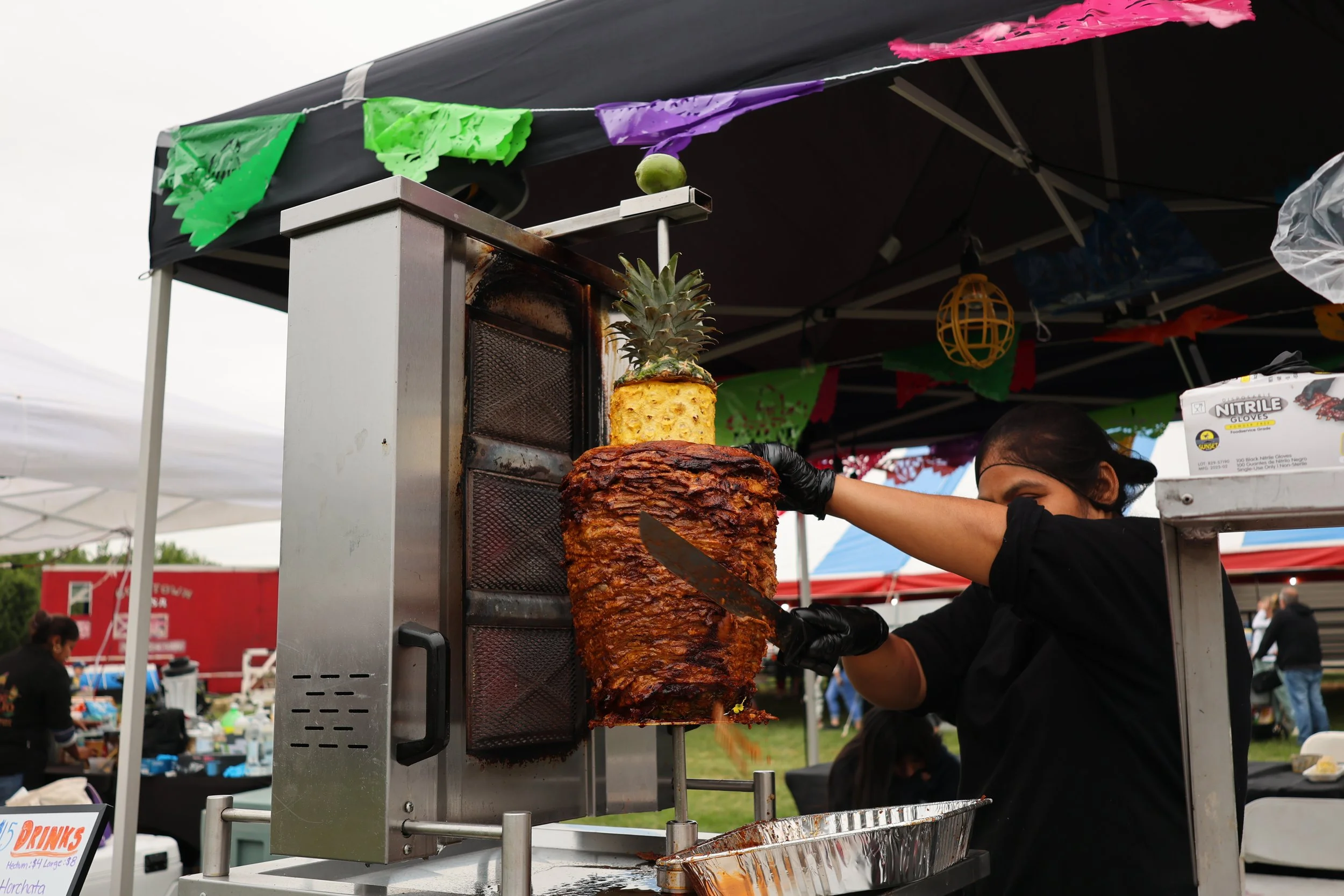 A vendor slices meat from a large vertical rotisserie at an outdoor fair or market. The rotisserie is topped with a pineapple. Colorful decorations and tents are visible in the background.
