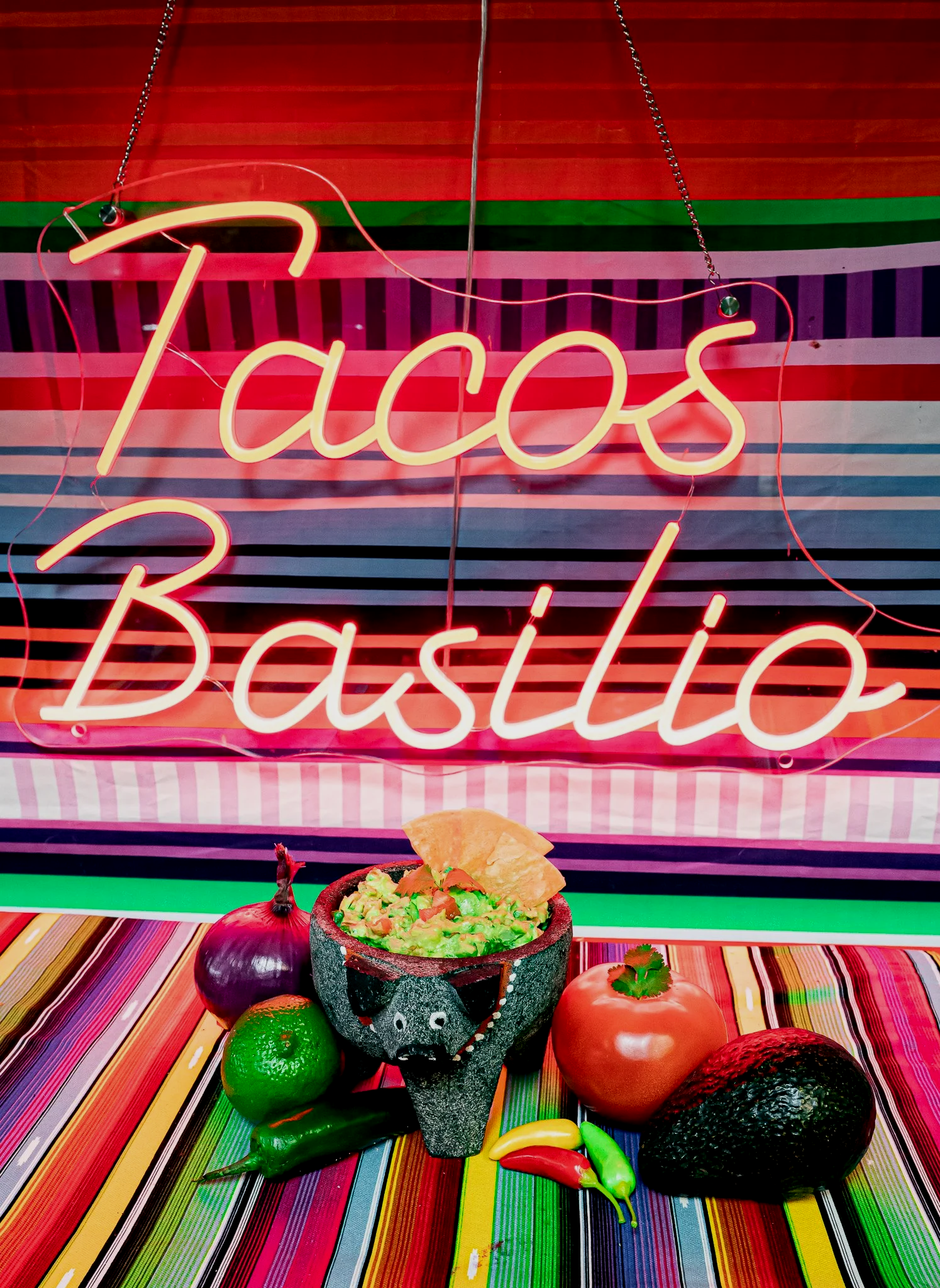 Colorful Mexican restaurant display with a neon sign reading 'Tacos' and 'Bachito', a bowl of guacamole with tortilla chips, and various vegetables including tomato, avocado, lime, and chili peppers on a multicolored striped tablecloth.