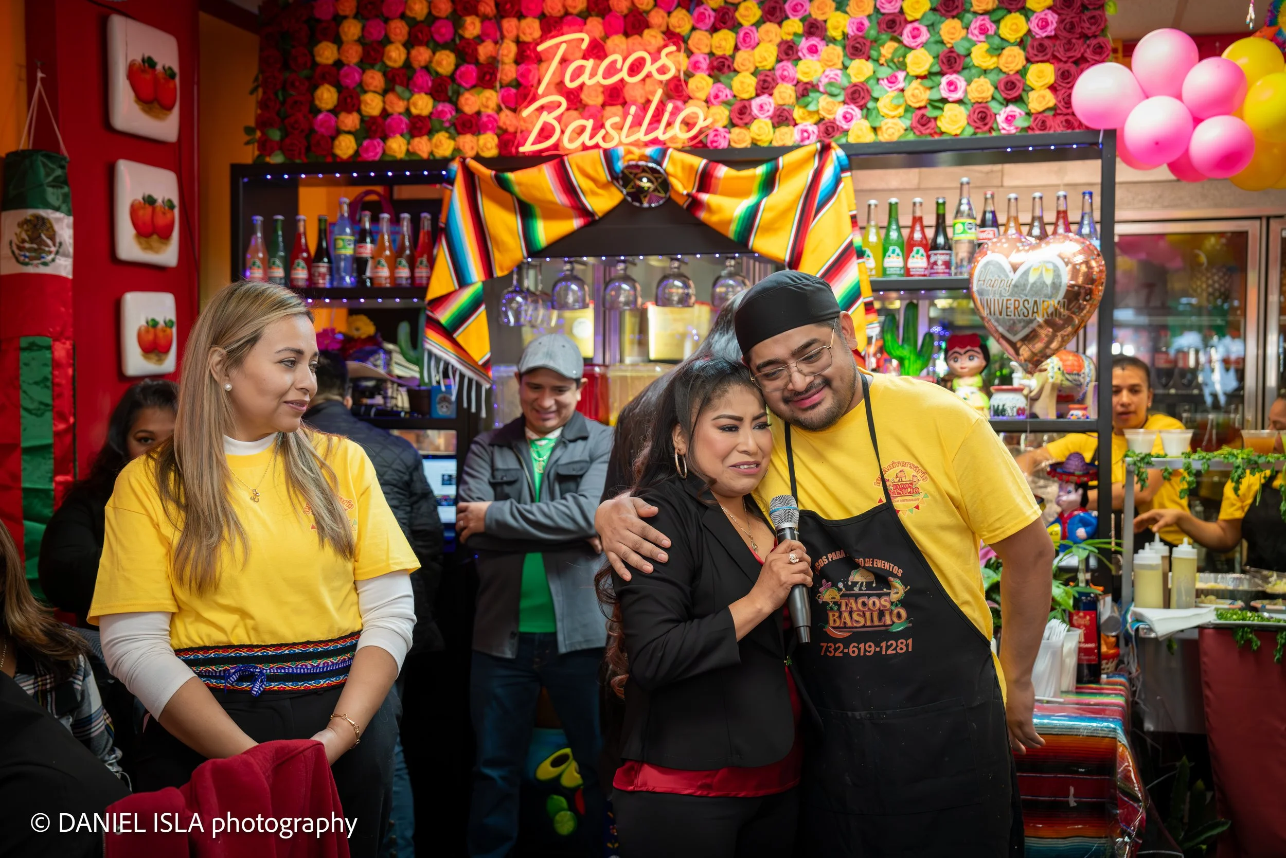 People gathered at a vibrant Mexican-themed event booth decorated with colorful flowers and balloons, with a neon sign reading 'Tacos Basilio.' A woman with a microphone is hugging a man in a yellow shirt and black cap, while others watch and smile i