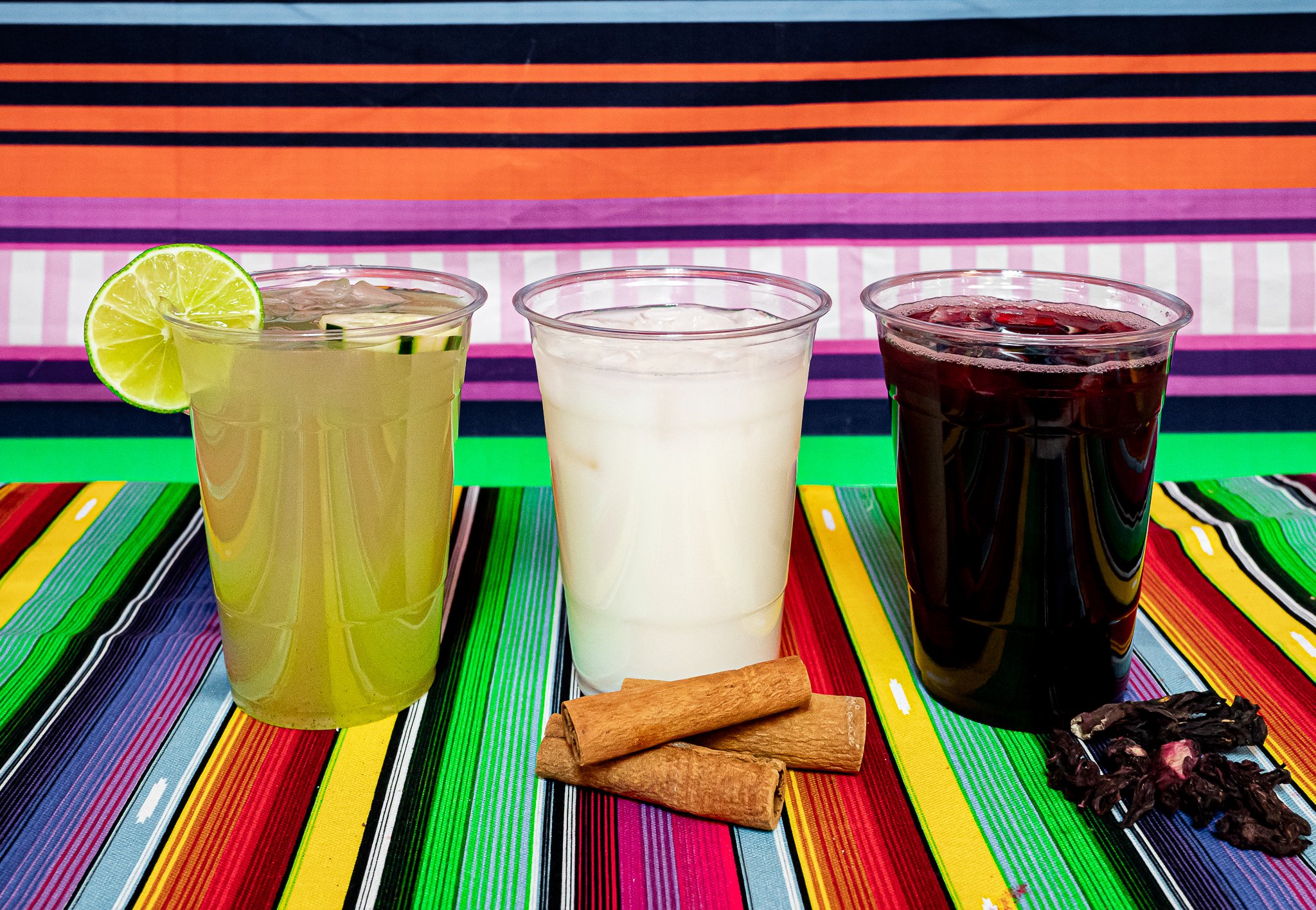 Three glasses filled with different colored drinks on a colorful striped tablecloth, with a lime wedge on the left glass, and some wafer and chocolate pieces in front.