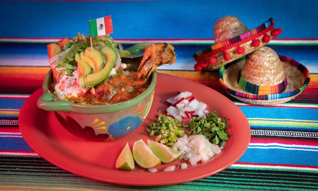 A colorful Mexican meal featuring a bowl of guacamole garnished with avocado slices and a small Mexican flag, accompanied by chopped onions, cilantro, and lime wedges on a terracotta plate, with a striped woven tablecloth and small sombreros in the b