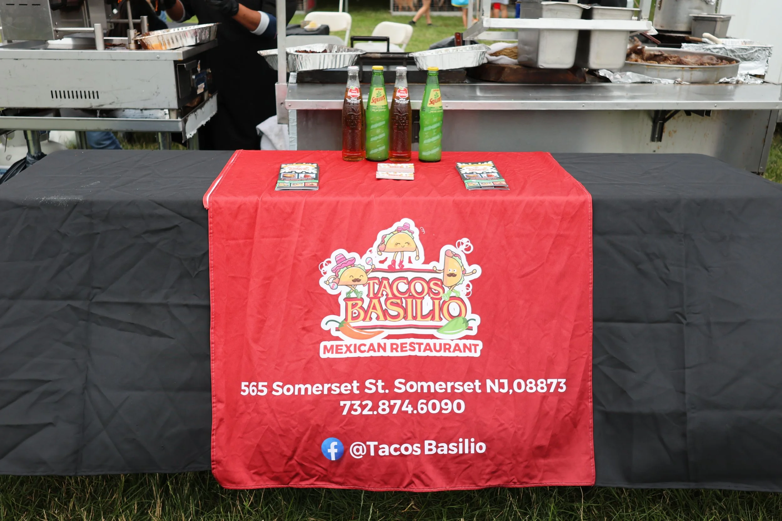 A table with a red tablecloth displaying the logo and contact information of Tacos Basilio, a Mexican restaurant, at an outdoor event. The background includes cooking equipment and people.