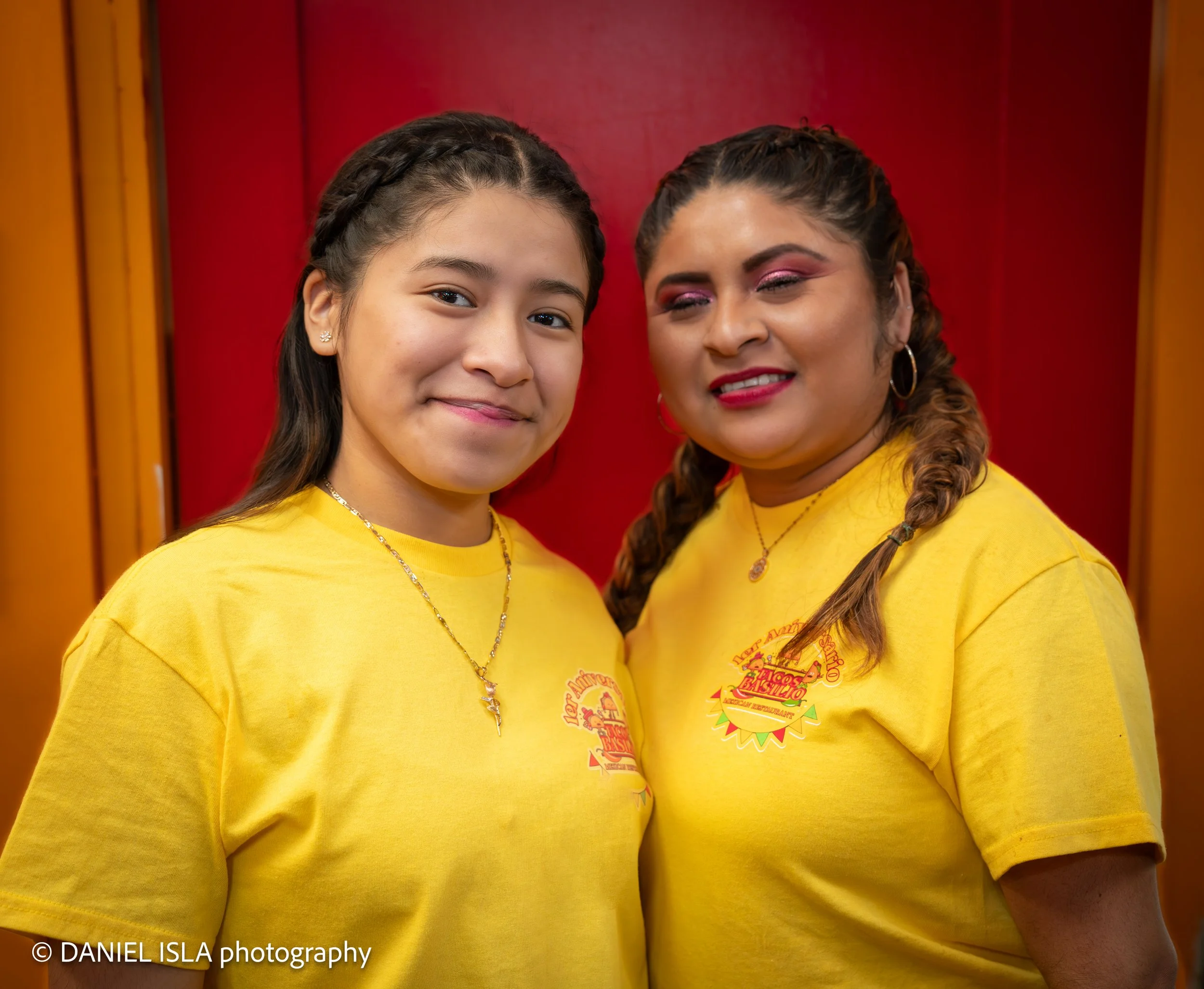 Two women wearing yellow T-shirts with a colorful logo, standing close together in front of a red background.