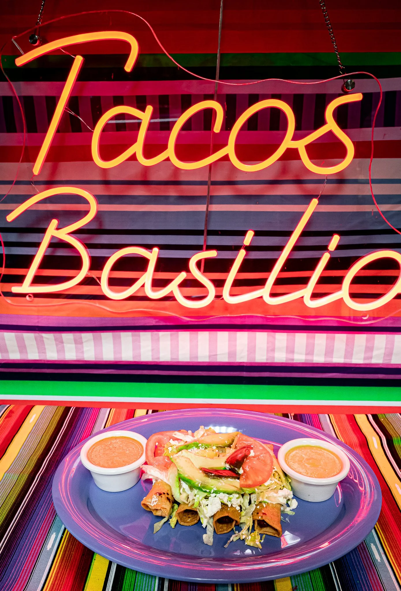 Colorful plate of Mexican food with tacos, guacamole, and salsa on a vibrant striped table under a neon sign that reads 'Tacos Basillo'.