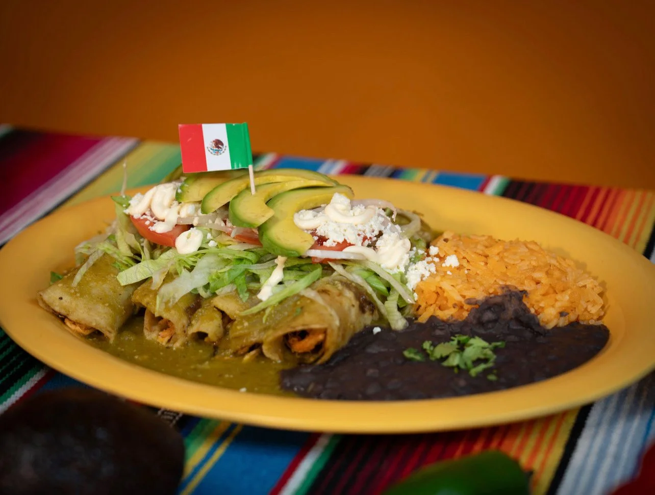 Plate of Mexican food with rice, beans, and tacos topped with avocado, tomato, lettuce, cheese, and a small Mexican flag.