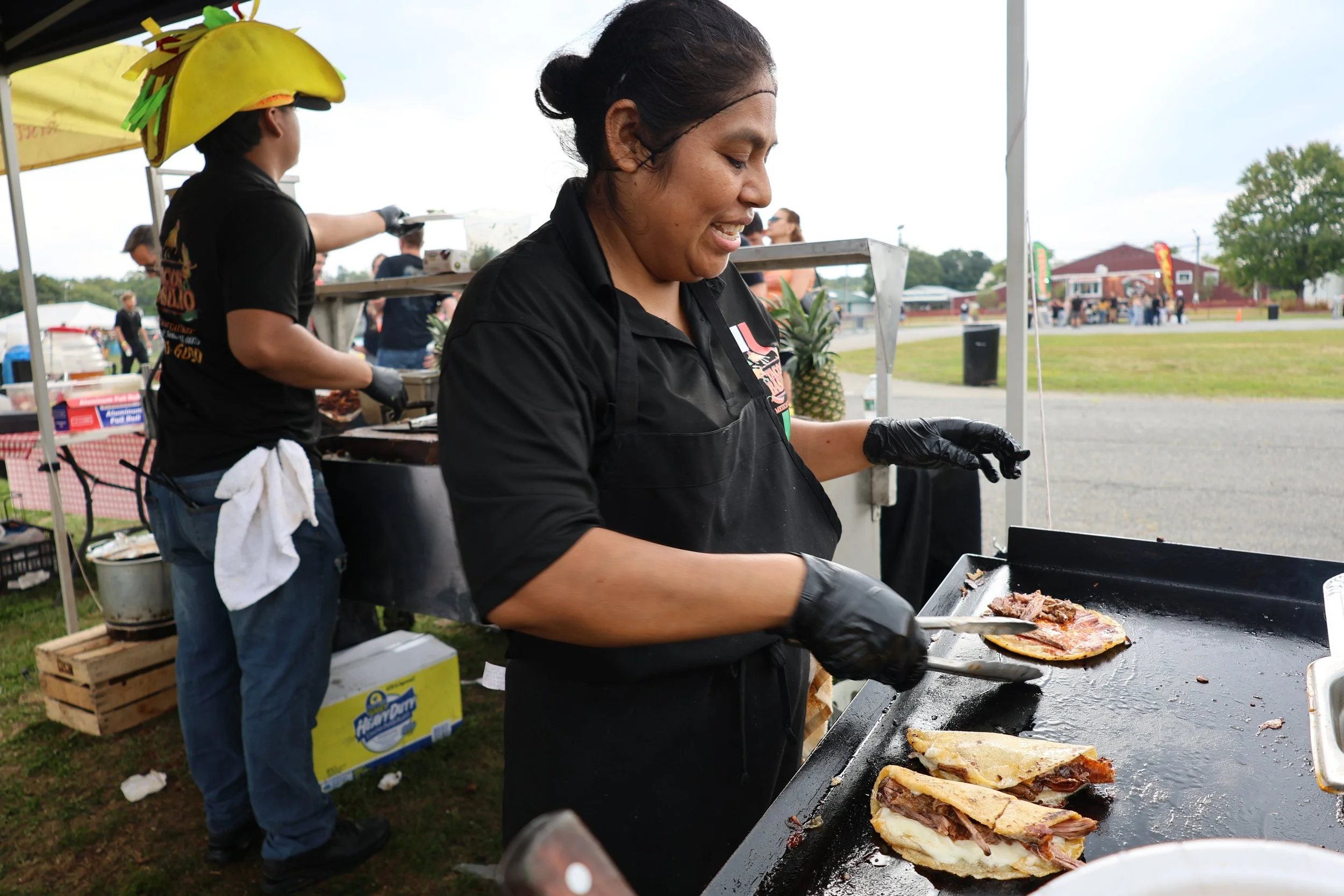 A woman wearing a black shirt and black gloves is cooking meat on a flat-top grill at an outdoor event. In the background, a person is wearing a taco-shaped hat with colorful paper decorations. There are people and tents visible in the distance on a 