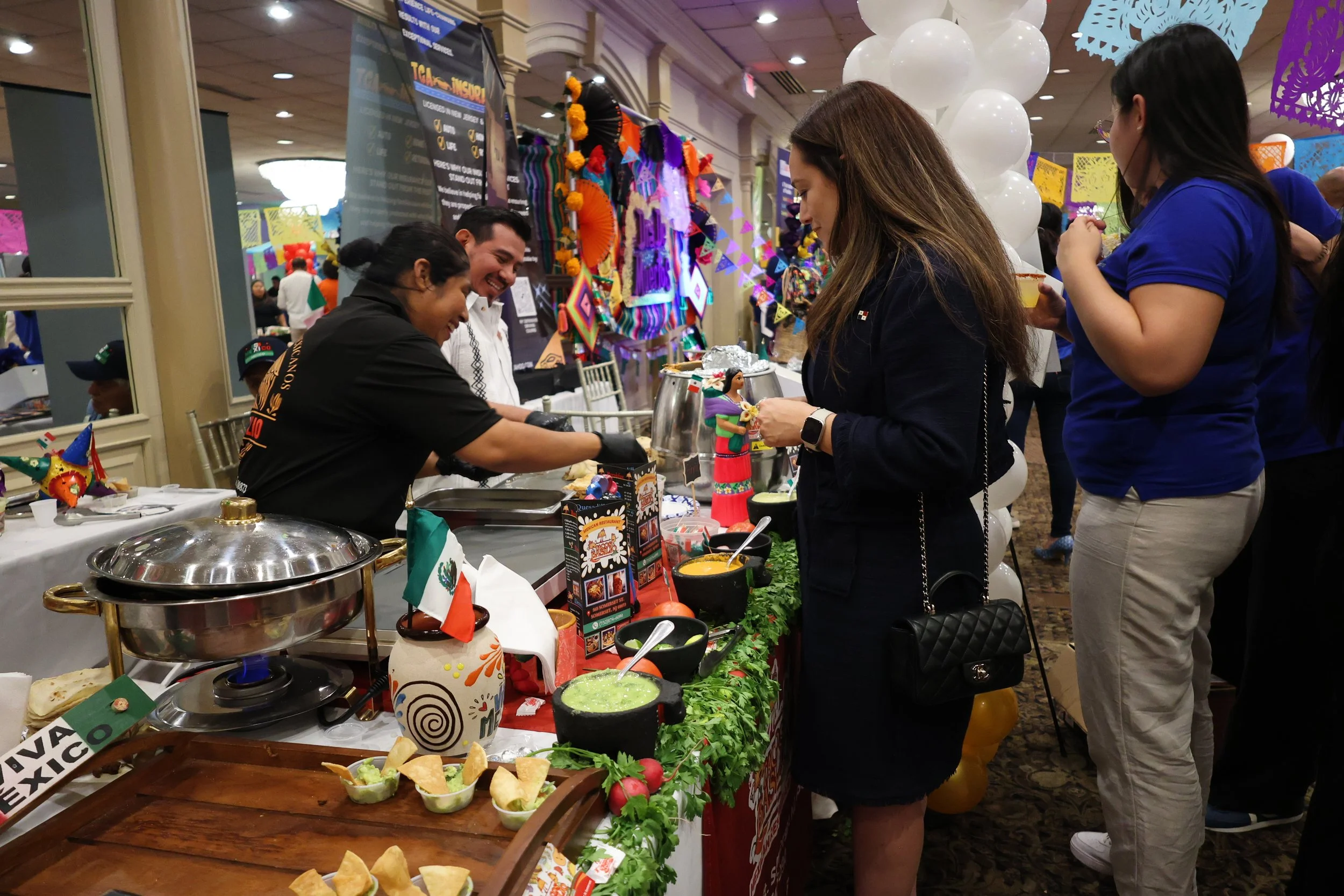 People enjoying a food and craft festival, with a table of Mexican food including tacos, guacamole, and decorative items, and colorful papel picado banners hanging overhead.