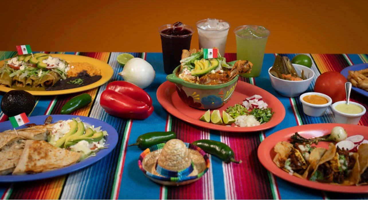 A colorful Mexican meal spread on a table with traditional striped tablecloths, including tacos, guacamole, salsas, chili, and beverages like iced soda and lime drink, garnished with hot peppers, lime wedges, and small flags of Mexico.