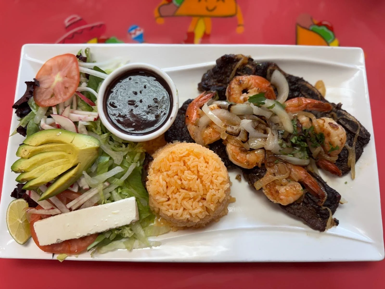 Plated Mexican meal with grilled steak topped with shrimp and onions, Mexican rice, side salad with tomato, avocado, radish, lettuce, and sliced lemon, and a small container of black sauce.