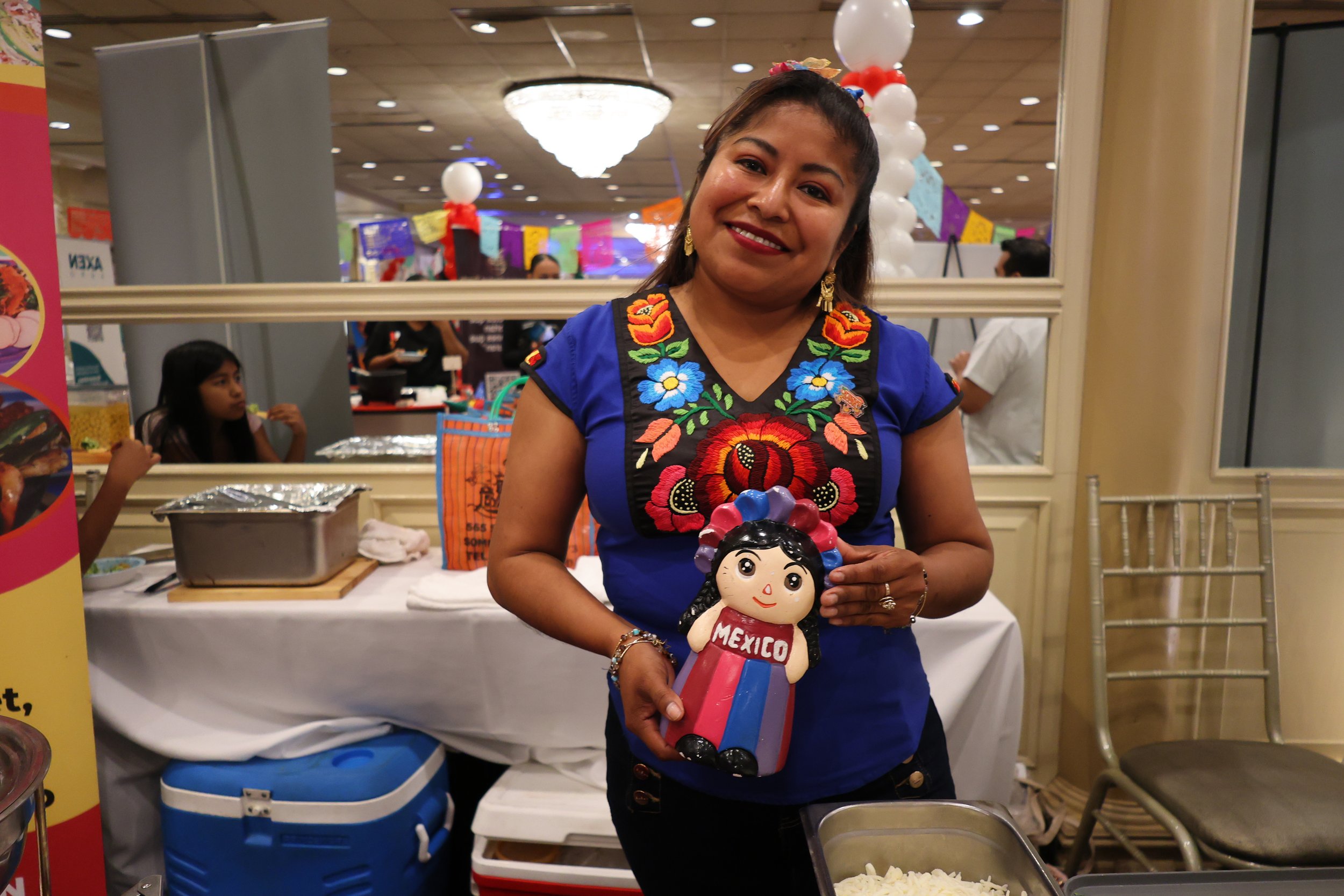 A woman wearing a vibrant embroidered blue dress stands inside a decorated event space, holding a colorful papel picado skeleton doll labeled 'Mexico', with balloons and festival decorations in the background.