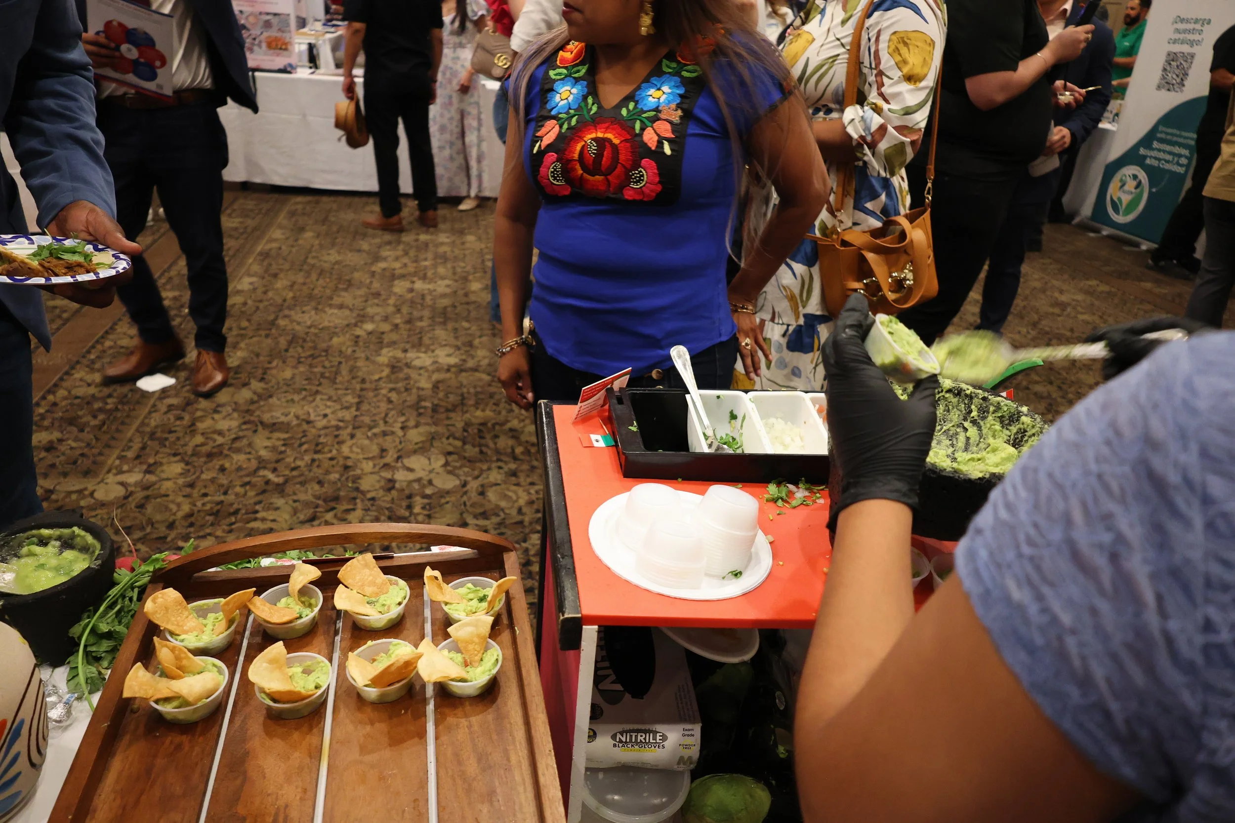 A group of people at a food event, with one person assembling small cups of guacamole topped with tortilla chips, and others in the background browsing booths. The setting appears to be an indoor expo or conference with a carpeted floor and vendor ta