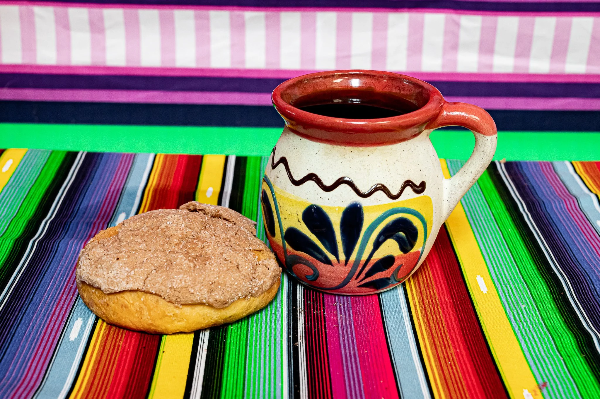 A colorful ceramic mug filled with black coffee, and a crumbly cookie on a vibrant striped tablecloth with pink, yellow, green, blue, black, and purple stripes.
