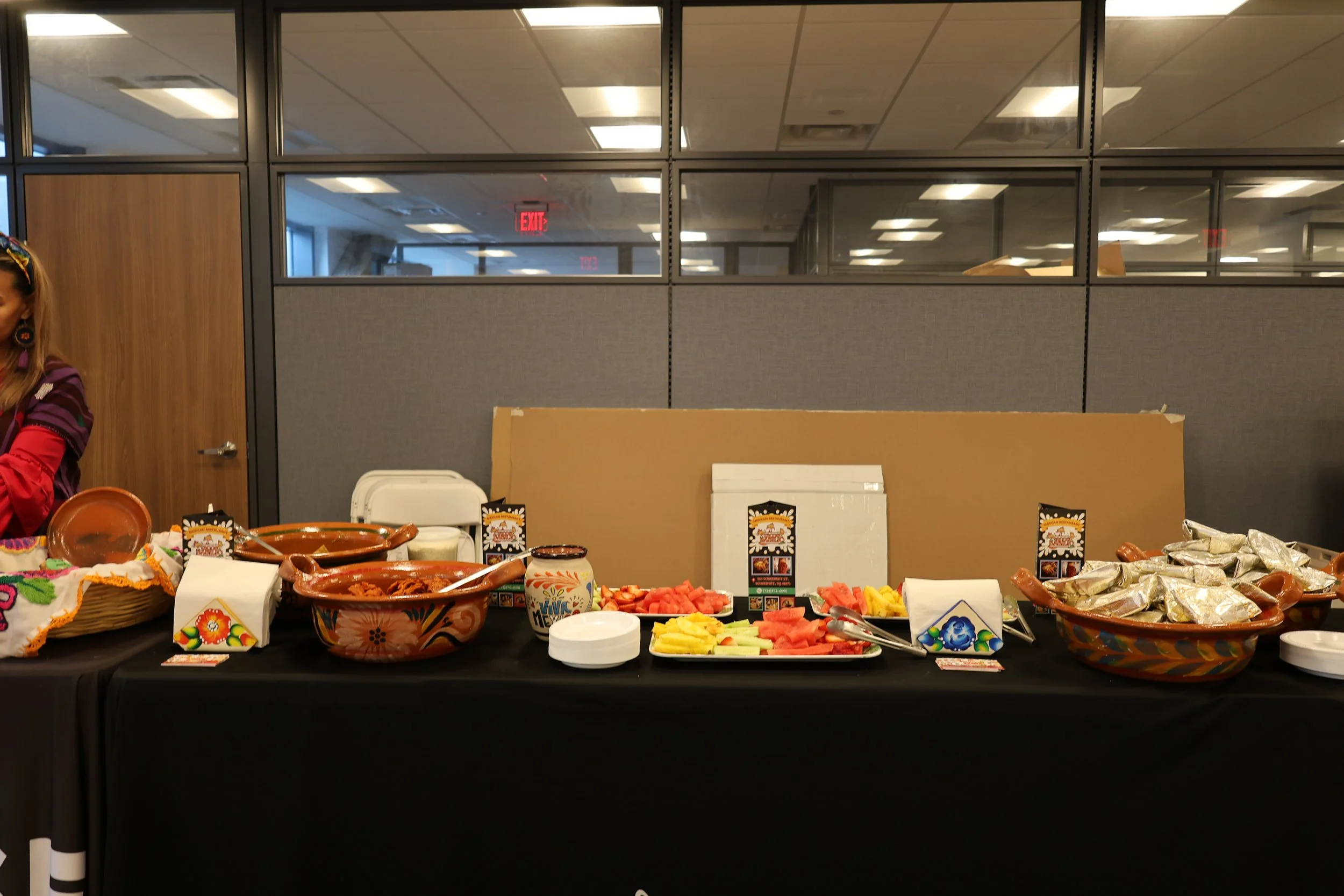 A table set up with a variety of Mexican food items including bowls of salsa, watermelon, and pineapple, with a person partially visible on the left side of the image and a background of office cubicles and ceiling lights.