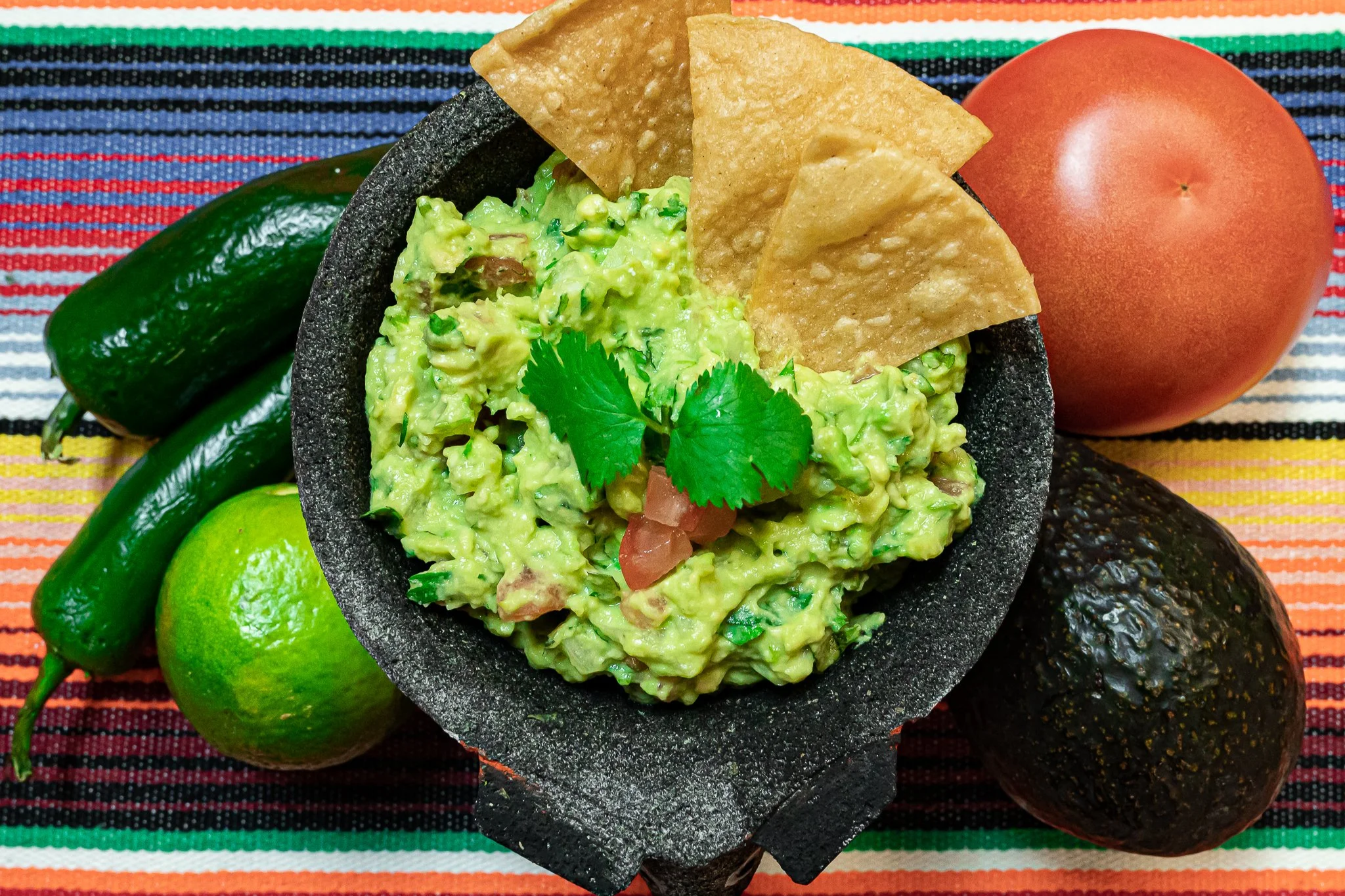 A bowl of guacamole with tortilla chips, a lime, a cucumber, a tomato, and an avocado on a colorful woven tablecloth.