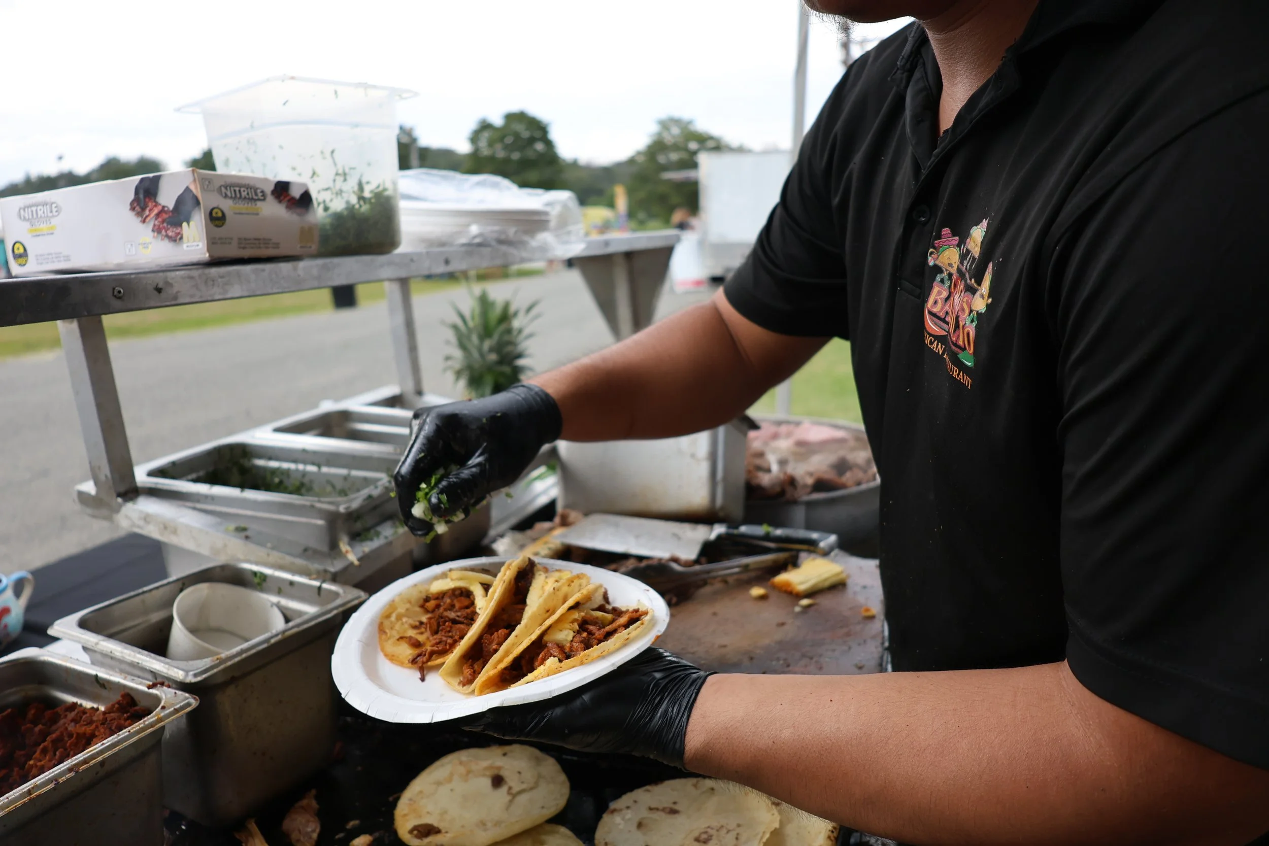 A person wearing a black shirt with a colorful logo and black gloves preparing Mexican street tacos with seasoned meat on a food cart.