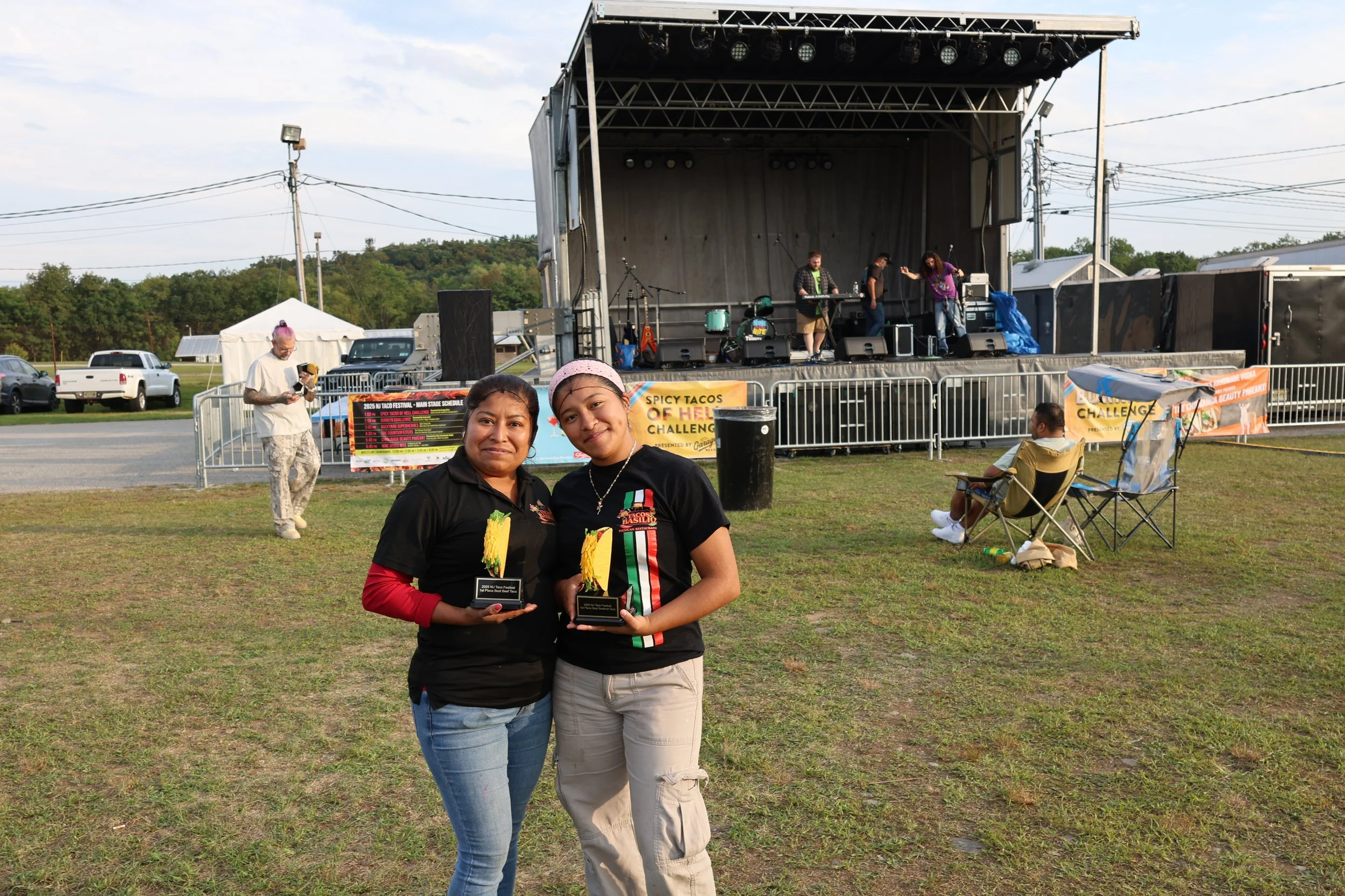Two women smiling and holding trophies at an outdoor festival, with a stage and other attendees in the background.