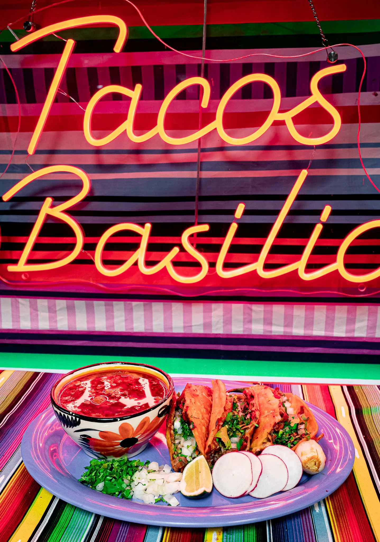 A colorful plate of Mexican food with tacos, radish slices, lemon wedges, chopped onions, cilantro, and a bowl of red sauce, on a multicolored striped table. A neon sign behind the plate reads 'Tacos Basilic'.
