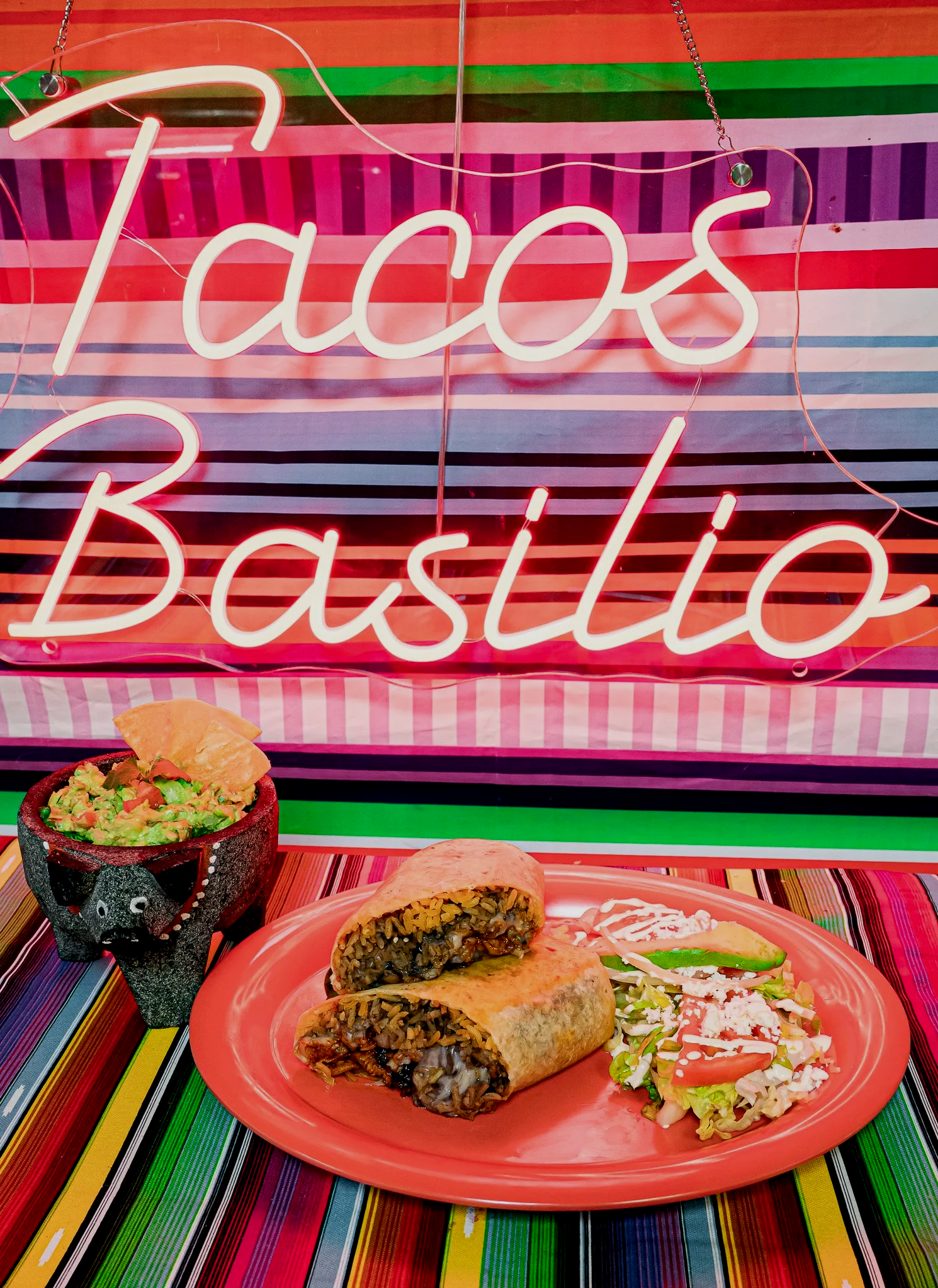 Colorful sign with pink neon lights that says 'Tacos Basilio.' Traditional Mexican food including tacos with seasoned meat, avocado garnish, and a side of fresh salad with tomatoes and shredded lettuce, served on a pink plate. The background features vibrant striped Mexican tablecloths.