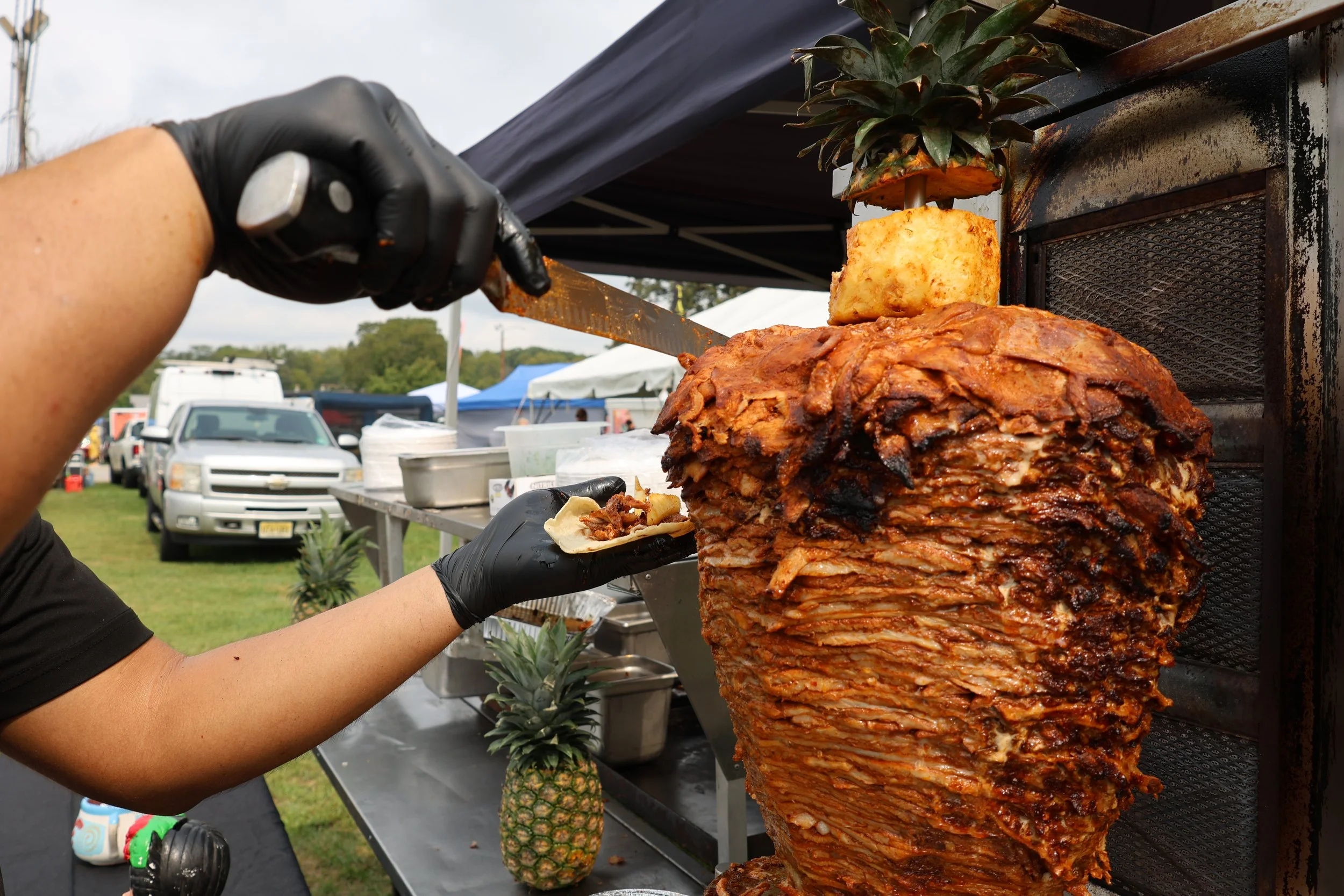 Person carving a large, cooked Mexican pork dish called 'lechón asado' on a vertical spit at an outdoor event.