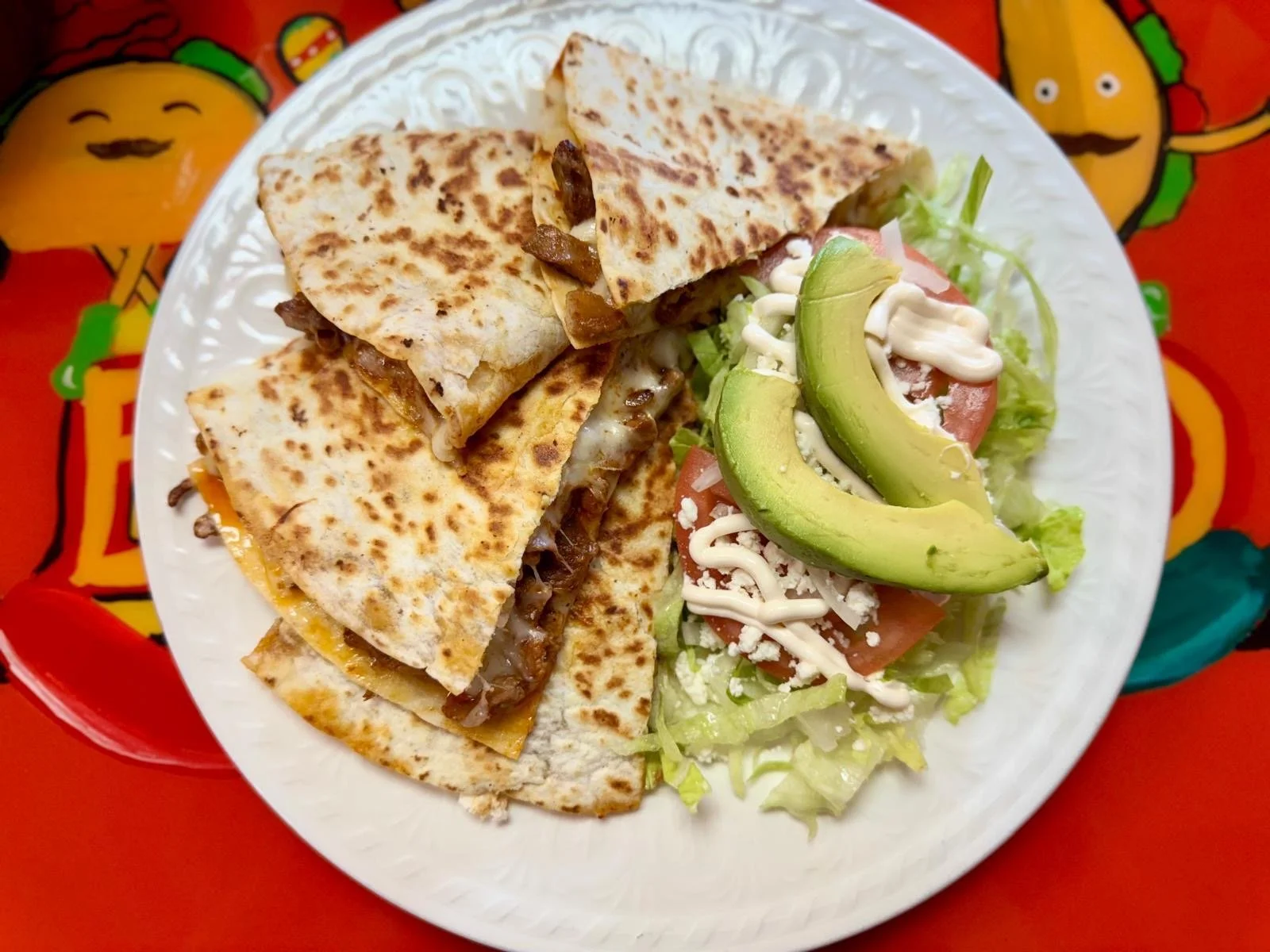 A plate of quesadillas with melted cheese and meat, served with a side salad that includes avocado slices, tomato, lettuce, shredded cheese, and mayonnaise, on a colorful background.