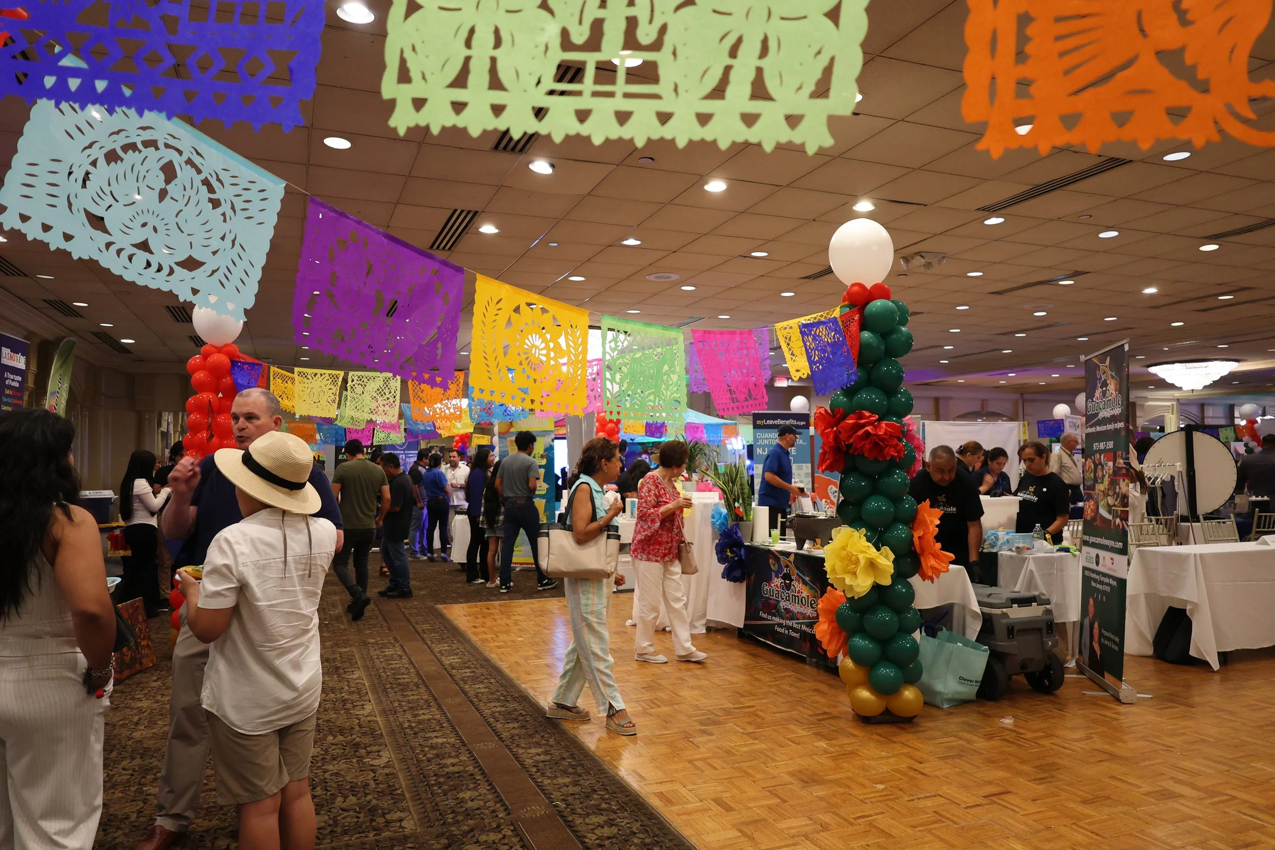 Colorful papel picado banners hanging overhead at a festive indoor event with various vendor booths and people shopping.