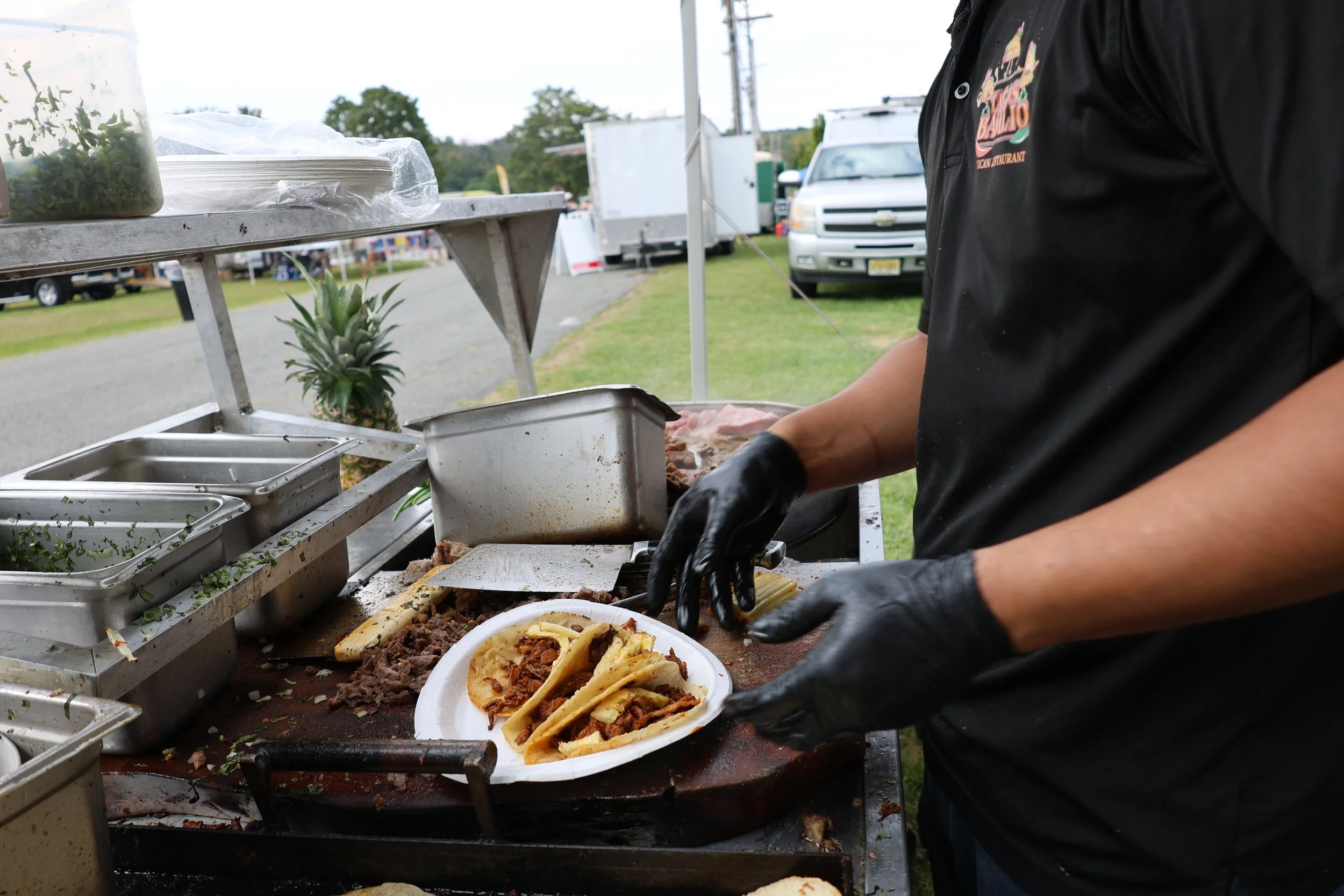 Person preparing tacos at an outdoor food stand, with various food containers and a pineapple on the table.