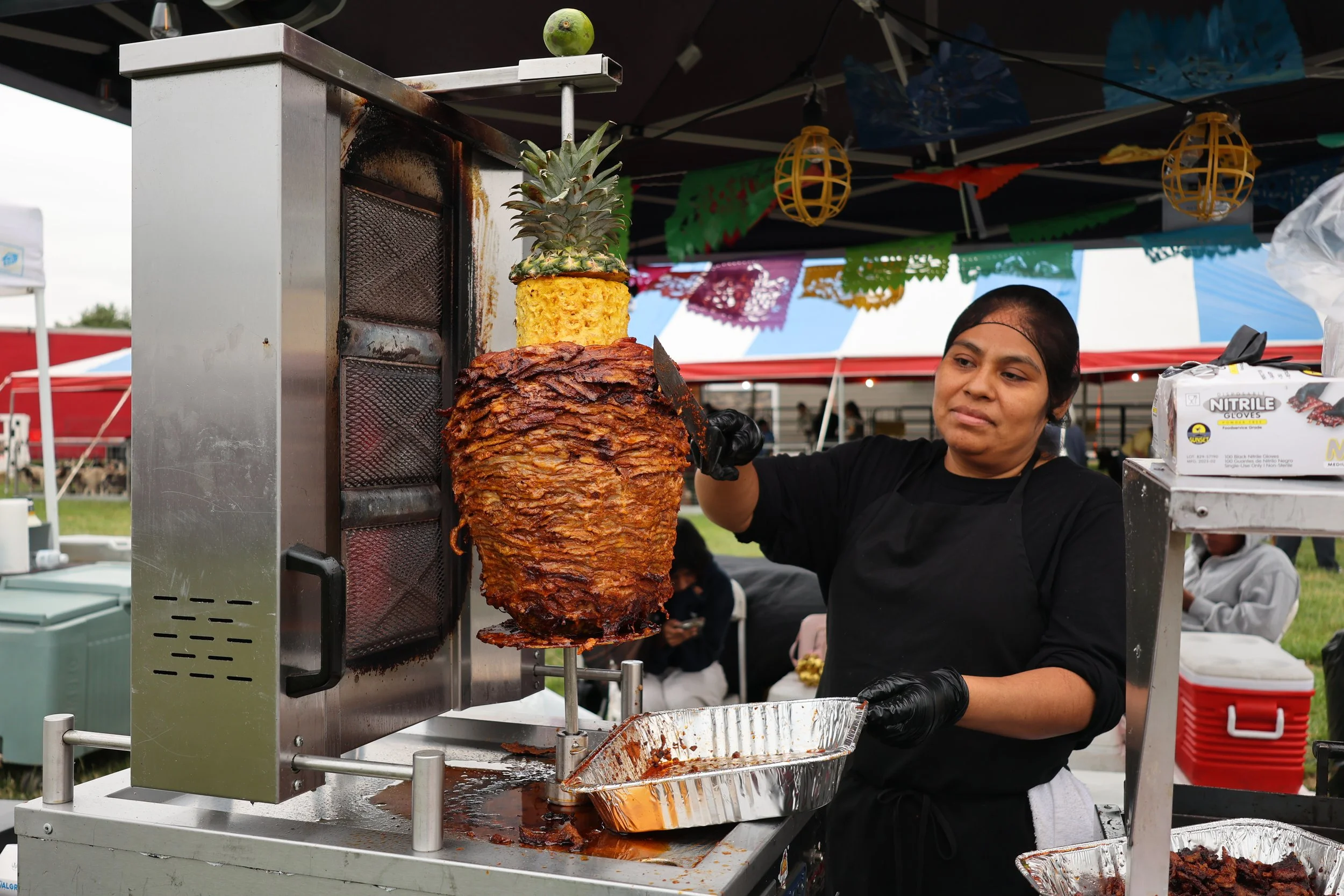 A woman wearing black gloves operating a vertical spit of roasted meat with a pineapple on top, at an outdoor event with colorful papel picado banners hanging above.