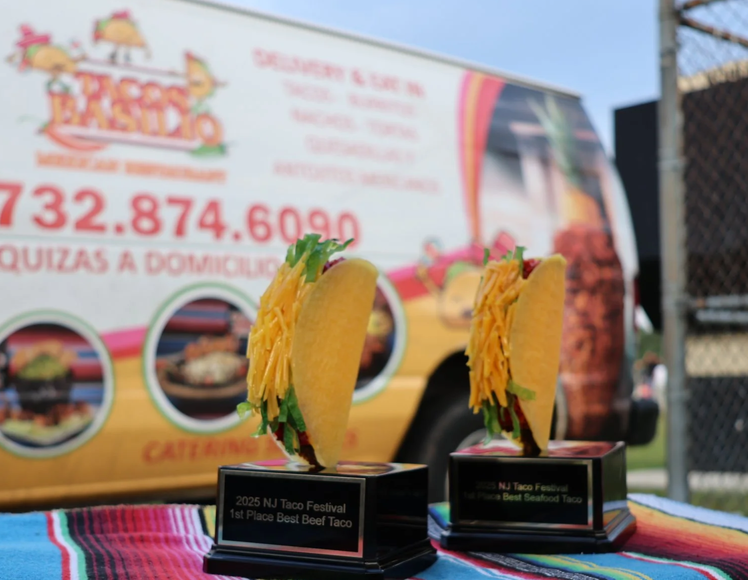 Two tacos with shredded cheese, lettuce, and beef on podiums at the 2023 New Jersey Taco Festival, with a food truck in the background advertising tacos and a phone number.