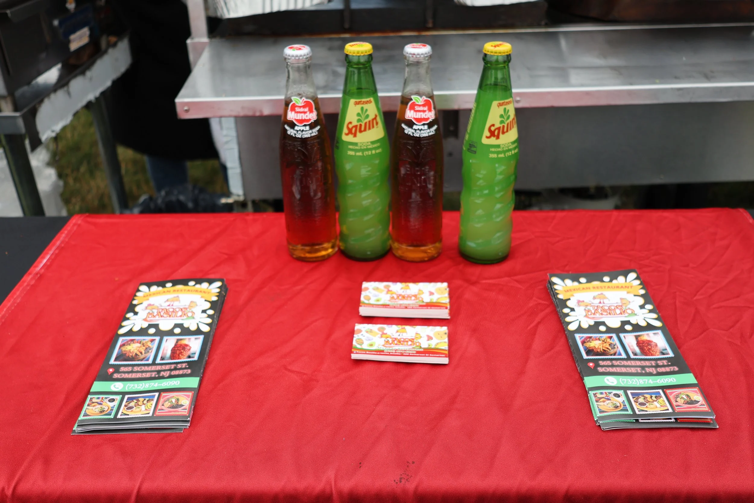 Four glass soda bottles on a red tablecloth with restaurant menus and stacked promotional cards, against a metallic background.