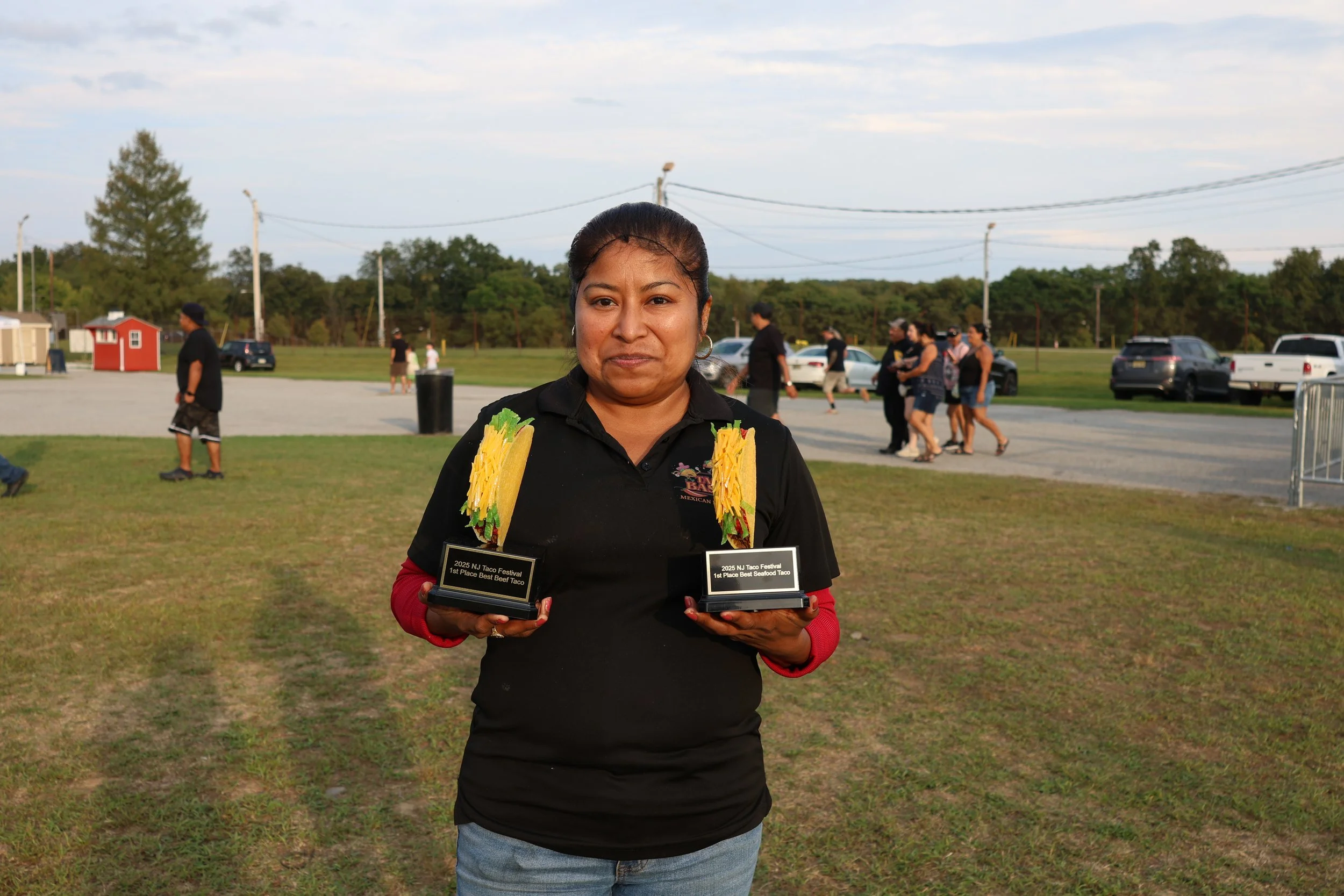 A woman holding two award plaques at an outdoor event with multiple people, cars, and trees in the background.