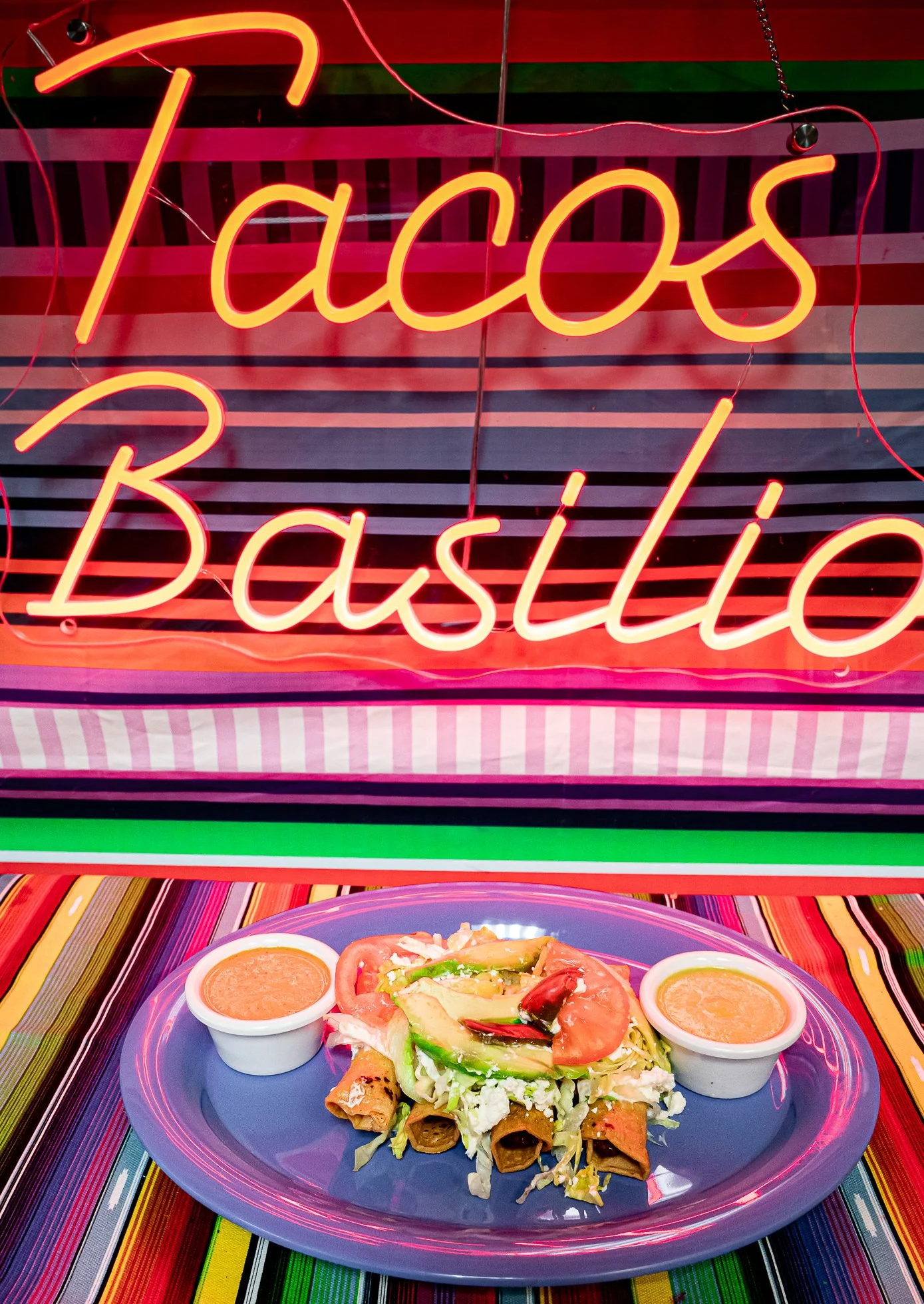 Display of a plate of Mexican food including tacos, with sides of sauce and garnishes, set on a colorful striped table with a neon sign that reads 'Tacos Basillo' in the background.