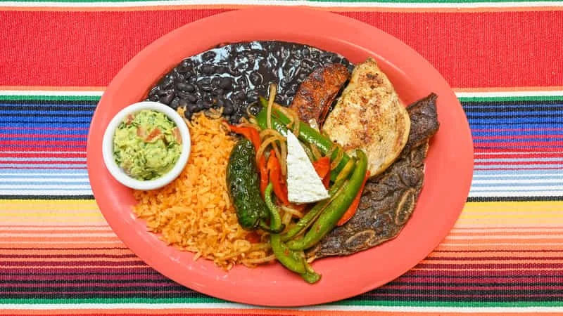 Mexican meal on a pink plate with rice, black beans, grilled chicken, grilled beef steak, guacamole, and colorful sautéed peppers and vegetables, on a striped colorful tablecloth.