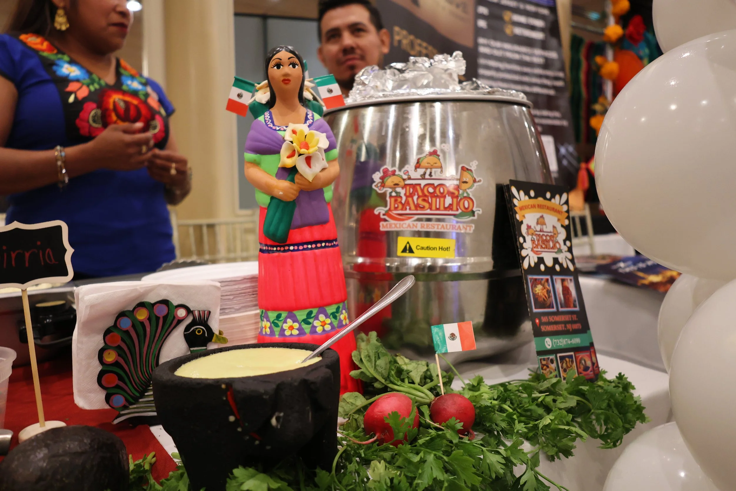 A colorful chef figurine wearing traditional Mexican attire, including a purple top, a green shawl, and a red skirt, standing behind a food display at a Mexican restaurant event. The display features fresh vegetables, herbs, and a pot with a yellow d