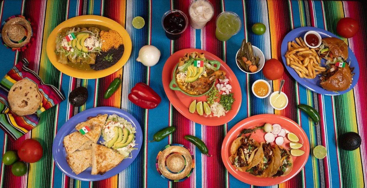 A colorful table with traditional Mexican food, including tacos, nachos, and enchiladas, along with drinks, tomatoes, jalapeños, limes, and condiments, all set on a vibrant Mexican textile background.