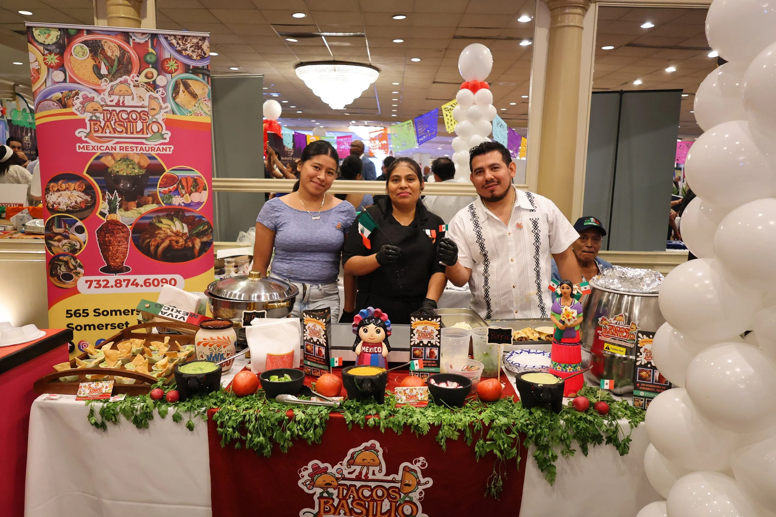 Three people standing behind a food stall at a Mexican restaurant event, with a colorful banner on the left and decorative balloons on the right, displaying various Mexican dishes and ingredients.