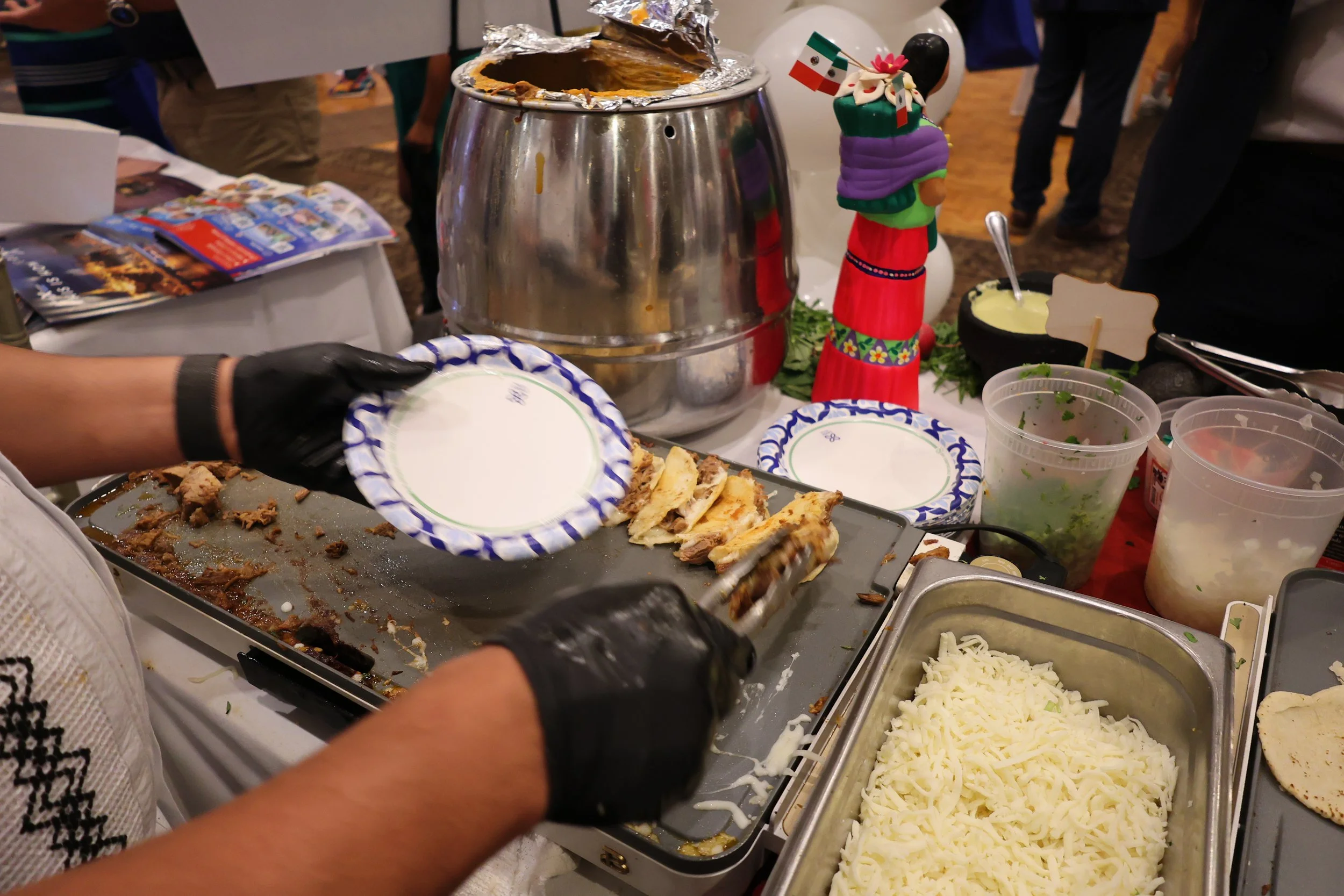 A table with various Mexican food items, including shredded cheese, grilled meat, and toppings. A person wearing black gloves is serving food from a tray. There is a hot container with a foil cover, a small Mexican flag, and decorative items on the t
