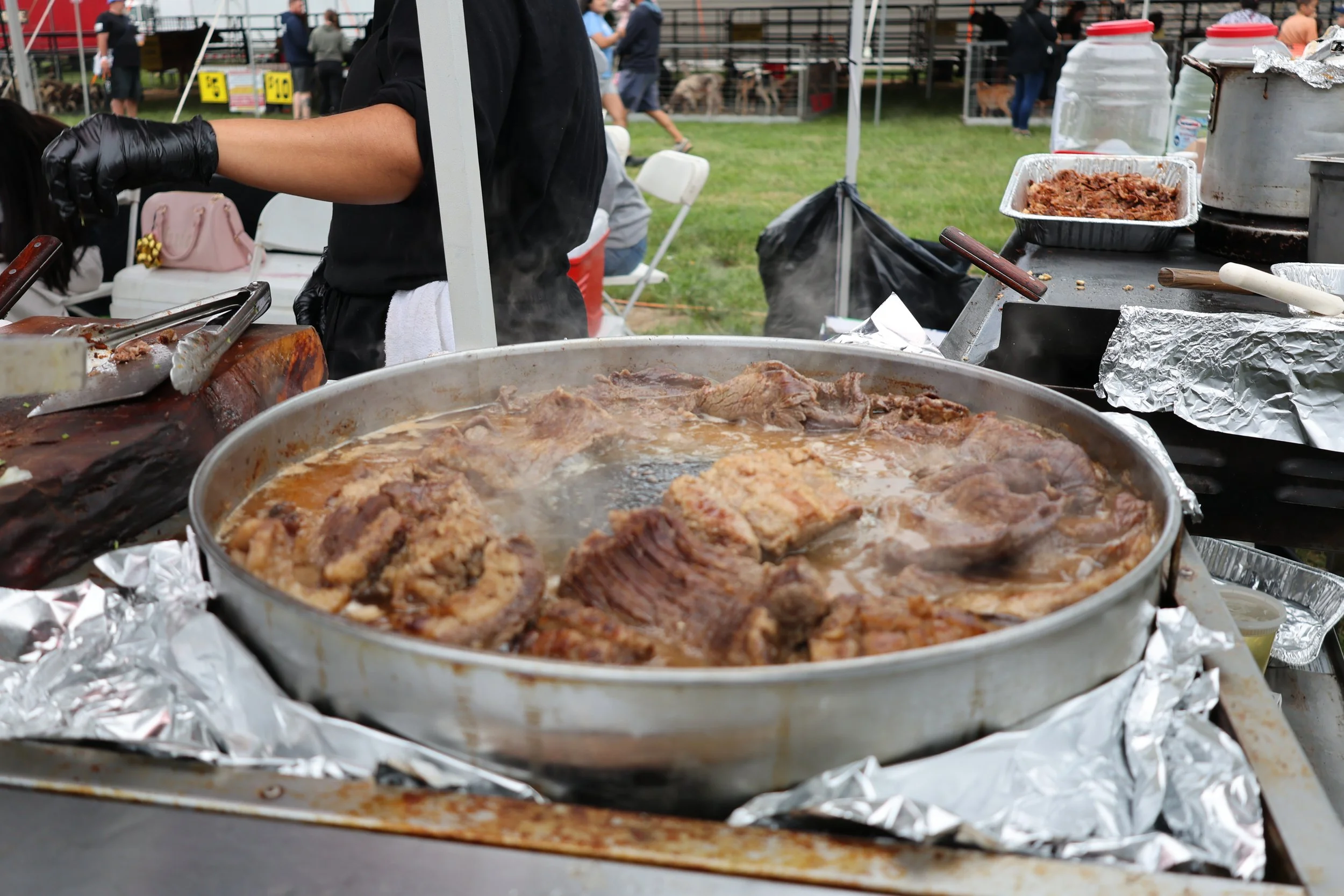 Outdoor food stall at a fair with large tray of cooked meat and foil-wrapped trays, people in the background